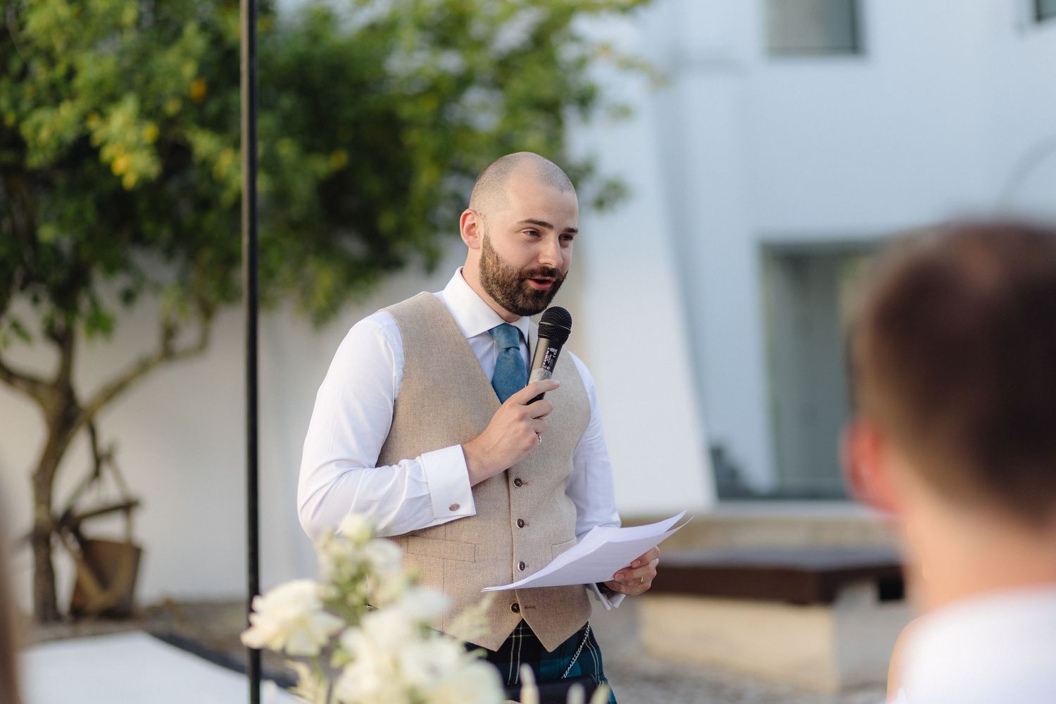 Guest speaking into microphone at outdoor reception with white building in background.