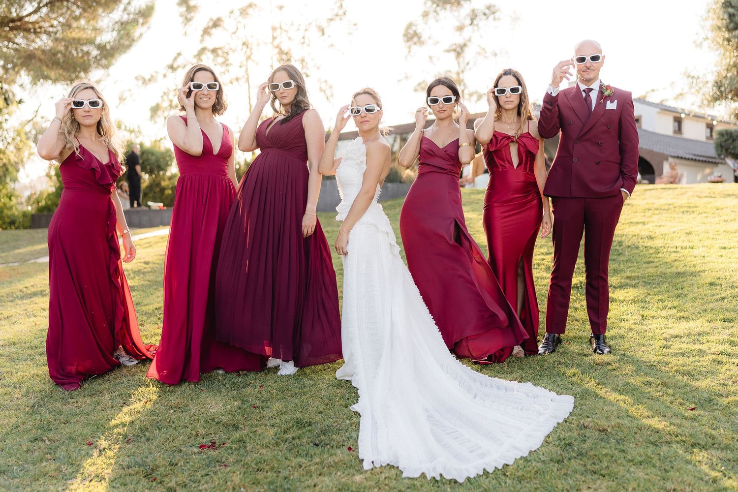 Wedding party in burgundy and white attire posing on grassy hill at sunset.