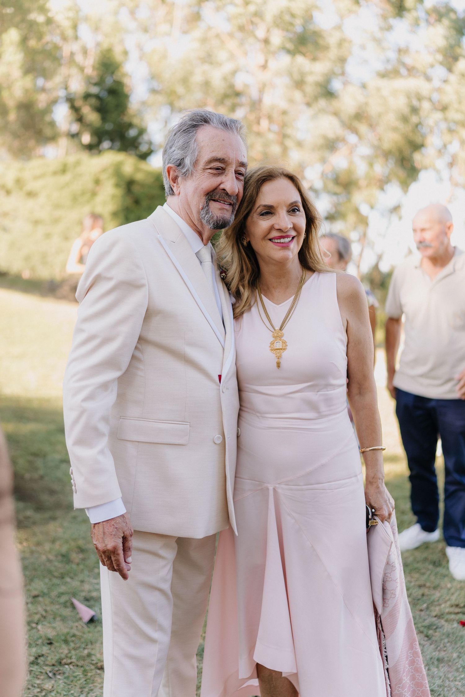 A couple wearing matching light colored formal attire poses together at an outdoor event.
