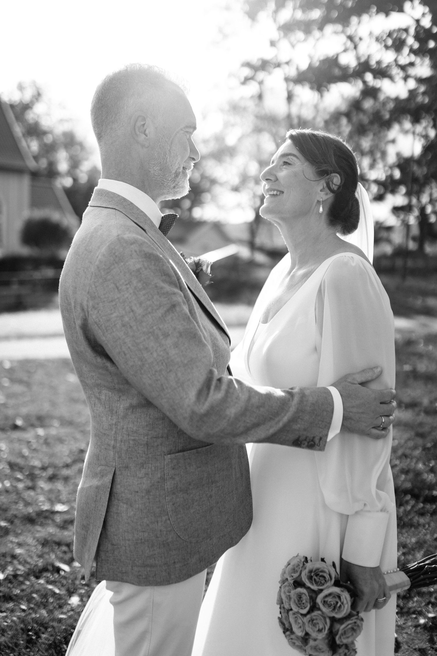 A romantic black and white portrait of newlyweds embracing outdoors in formal wedding attire.