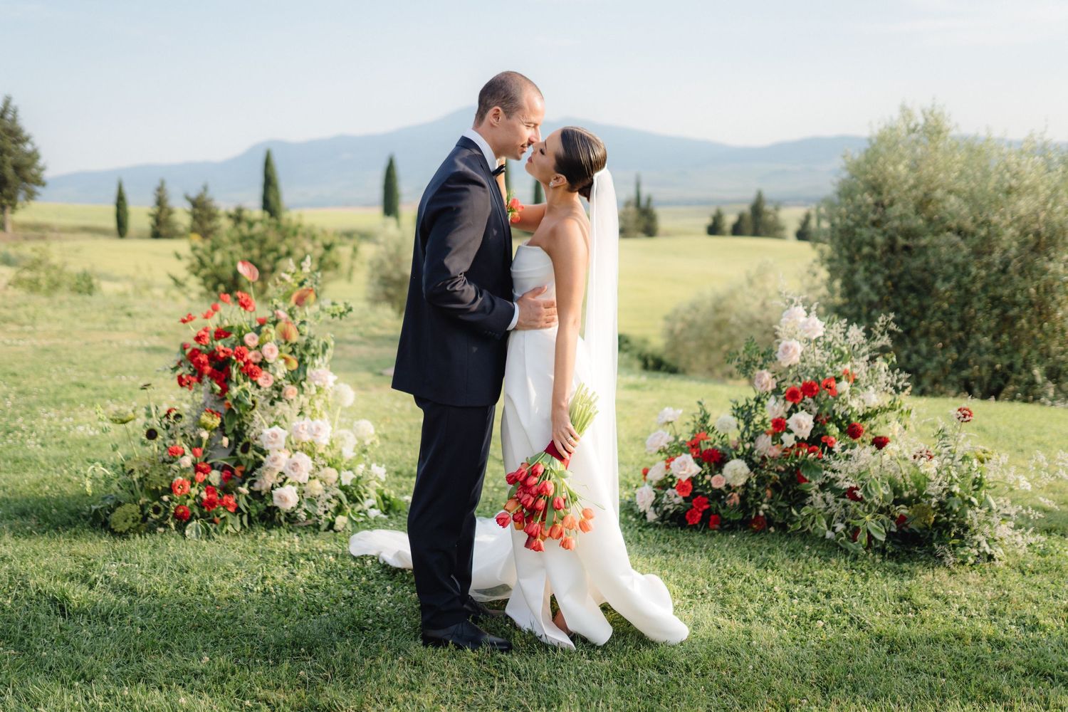 A couple shares a romantic kiss at an outdoor wedding ceremony surrounded by lush floral arrangements.