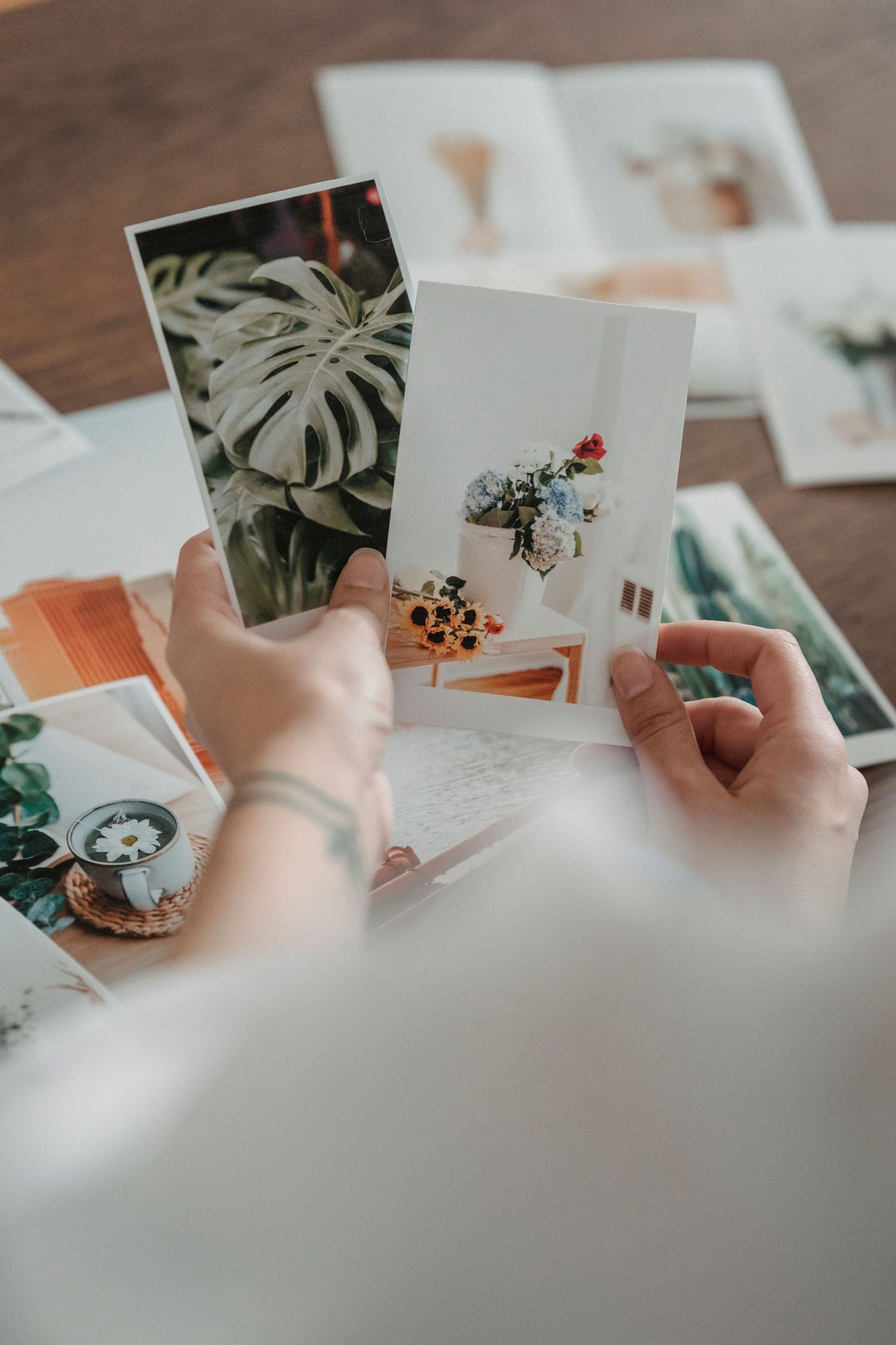Hands holding printed photos of tropical plants and flowers over a wooden table scattered with more botanical prints.