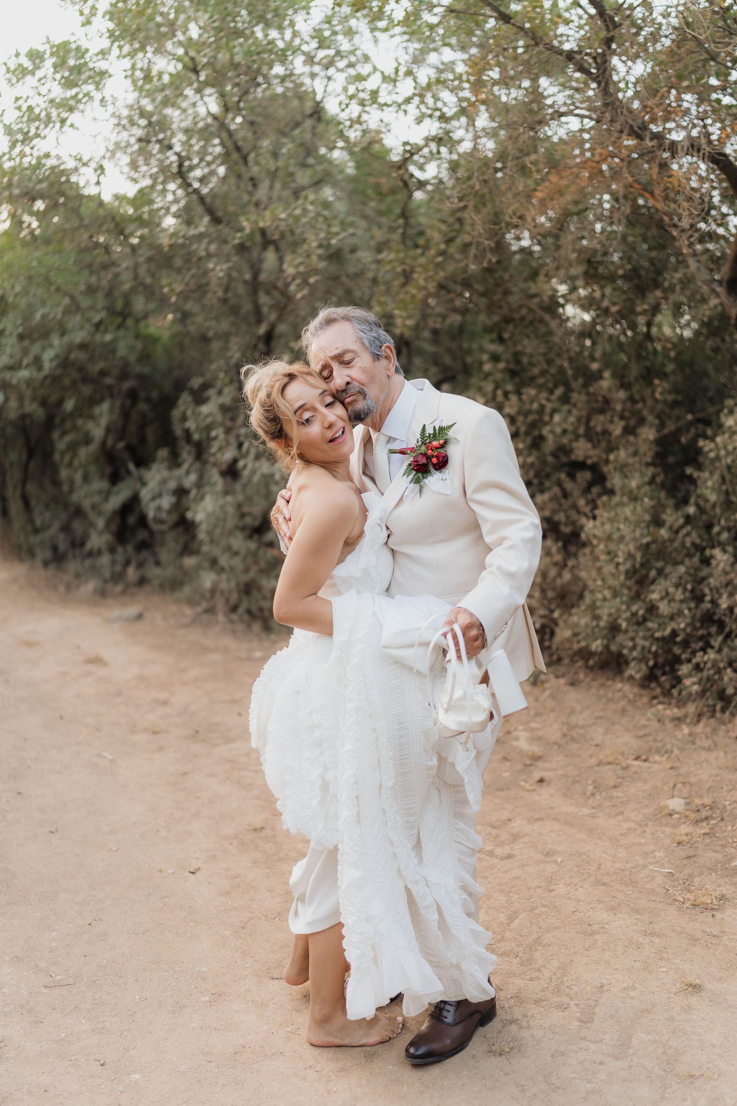 Newlyweds in white attire sharing intimate moment on outdoor trail.