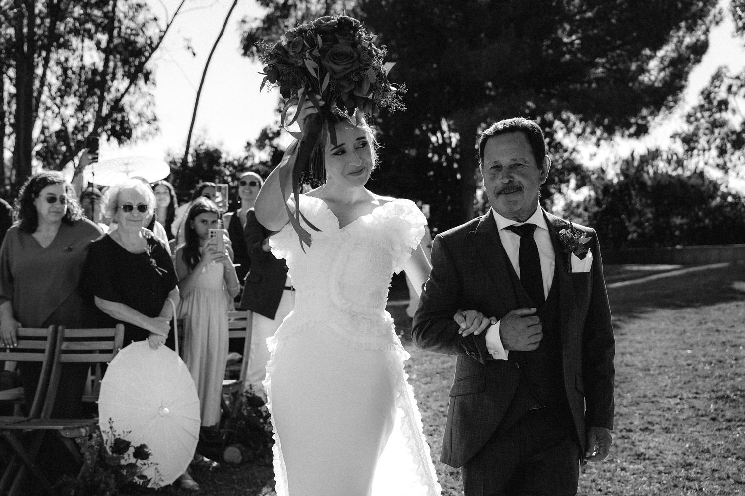 A black and white photo of a couple walking together during an outdoor wedding ceremony.