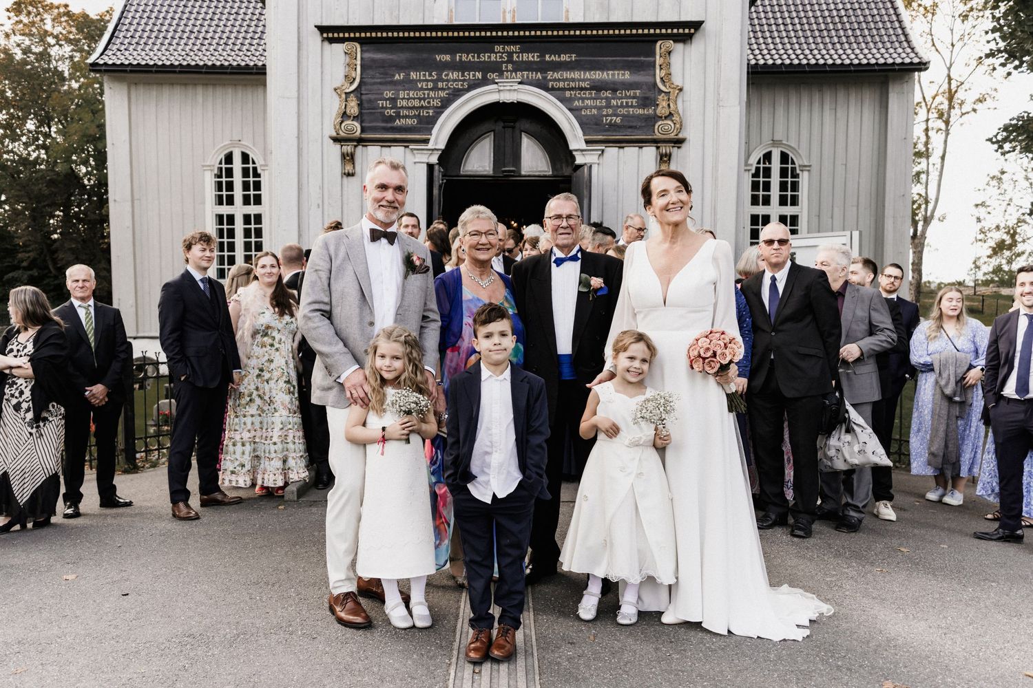 Wedding party and guests gathered outside a traditional white church building.