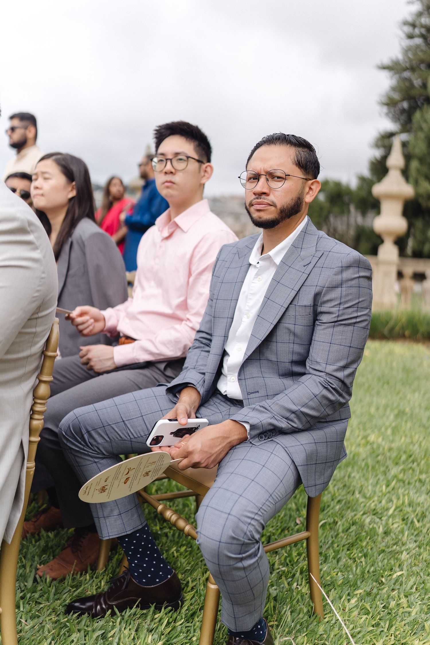 Wedding guests in formal attire seated on wooden chairs during an outdoor ceremony.
