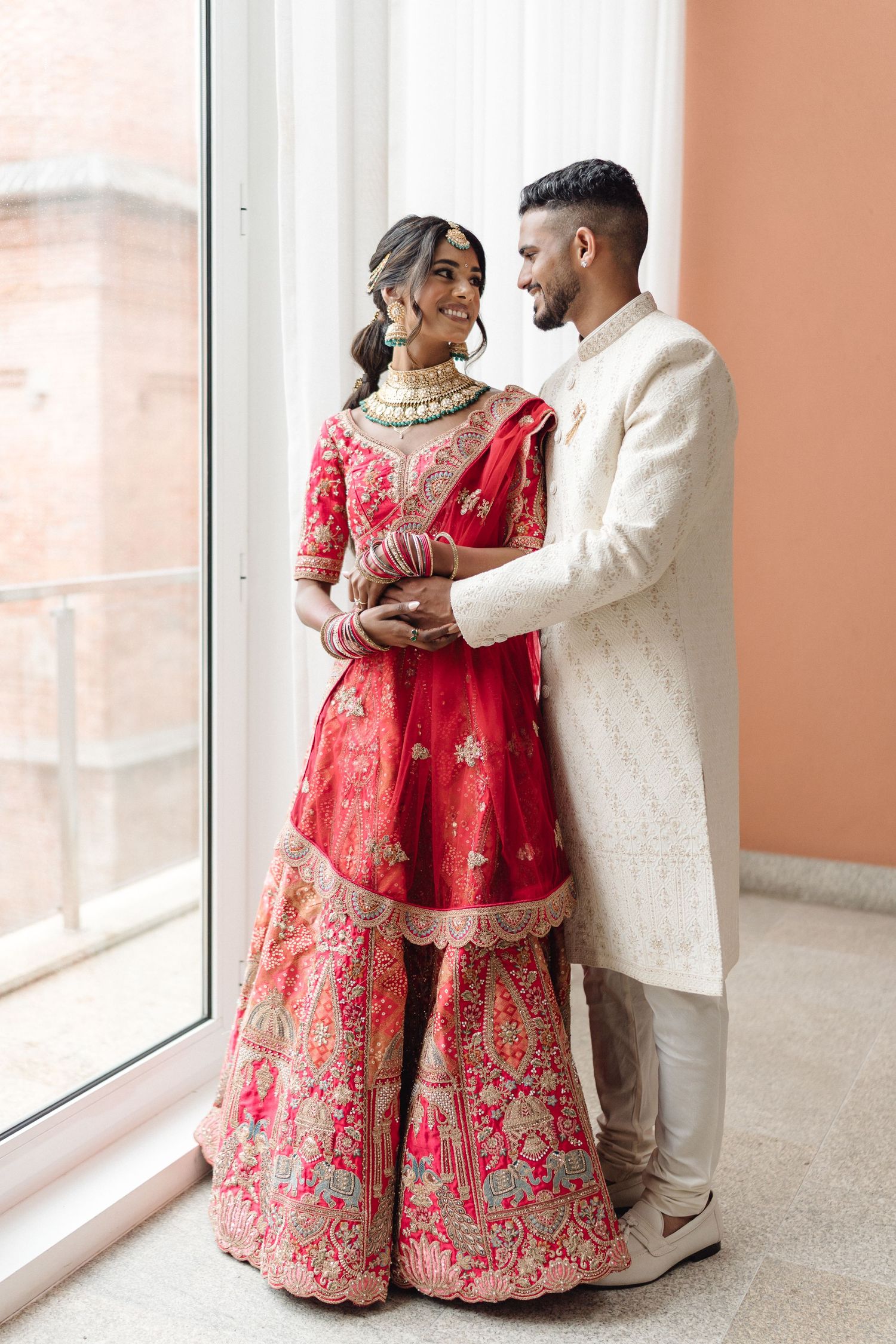 A couple in traditional South Asian wedding attire posing by bright windows.
