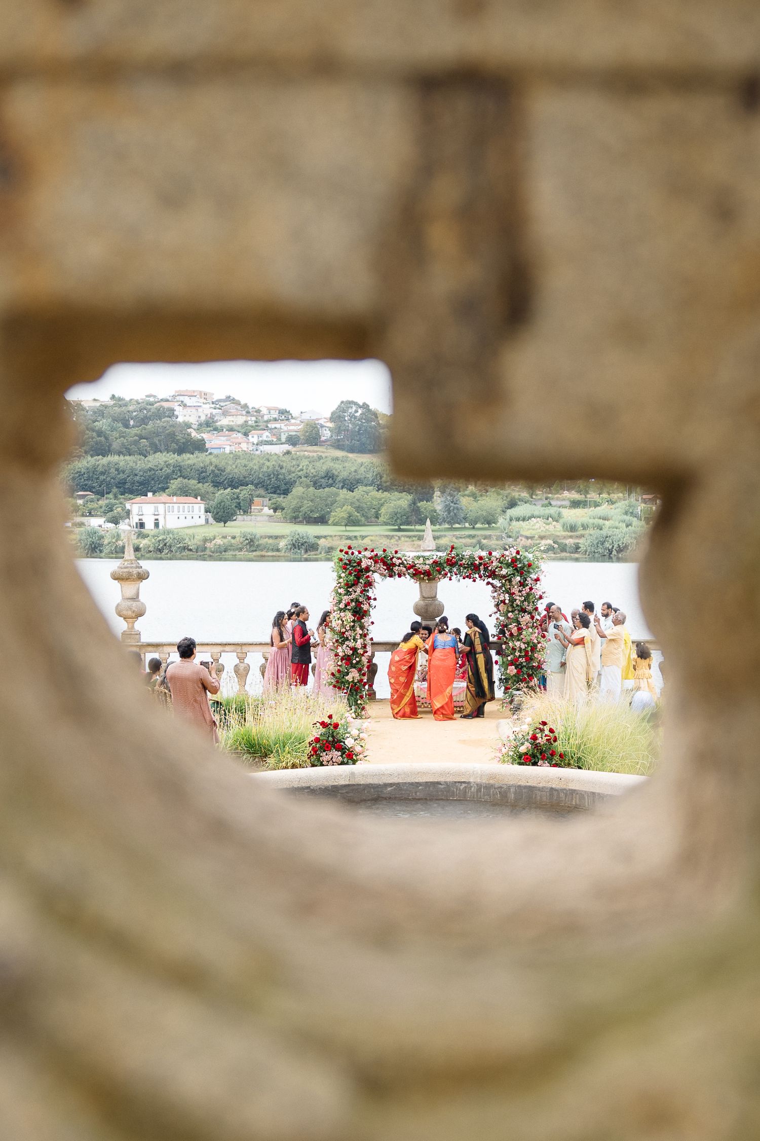 Wedding ceremony setup viewed through stone architecture with floral decorations and water view.