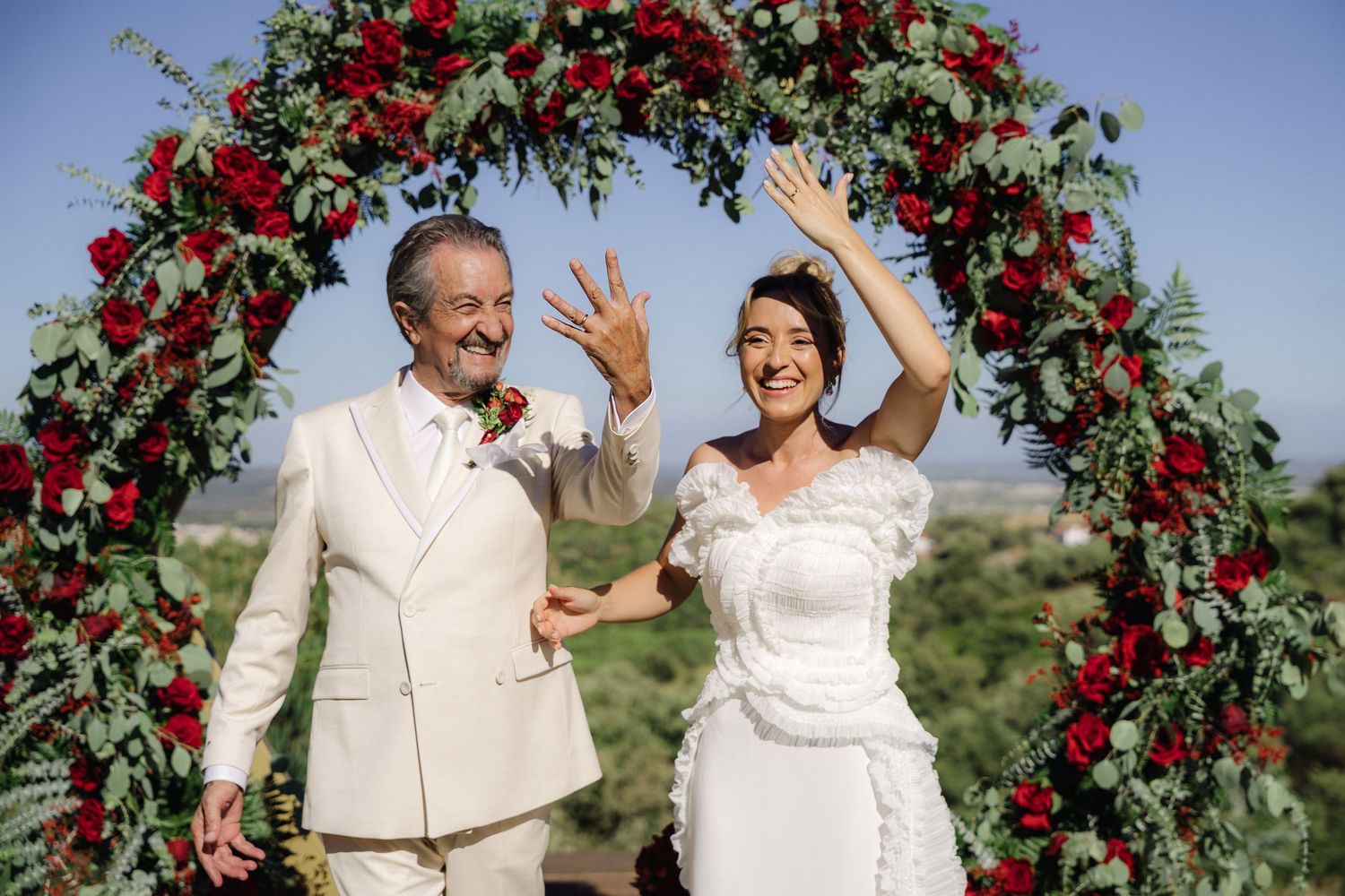 Celebratory poses under a red rose arch against blue sky during outdoor wedding ceremony.