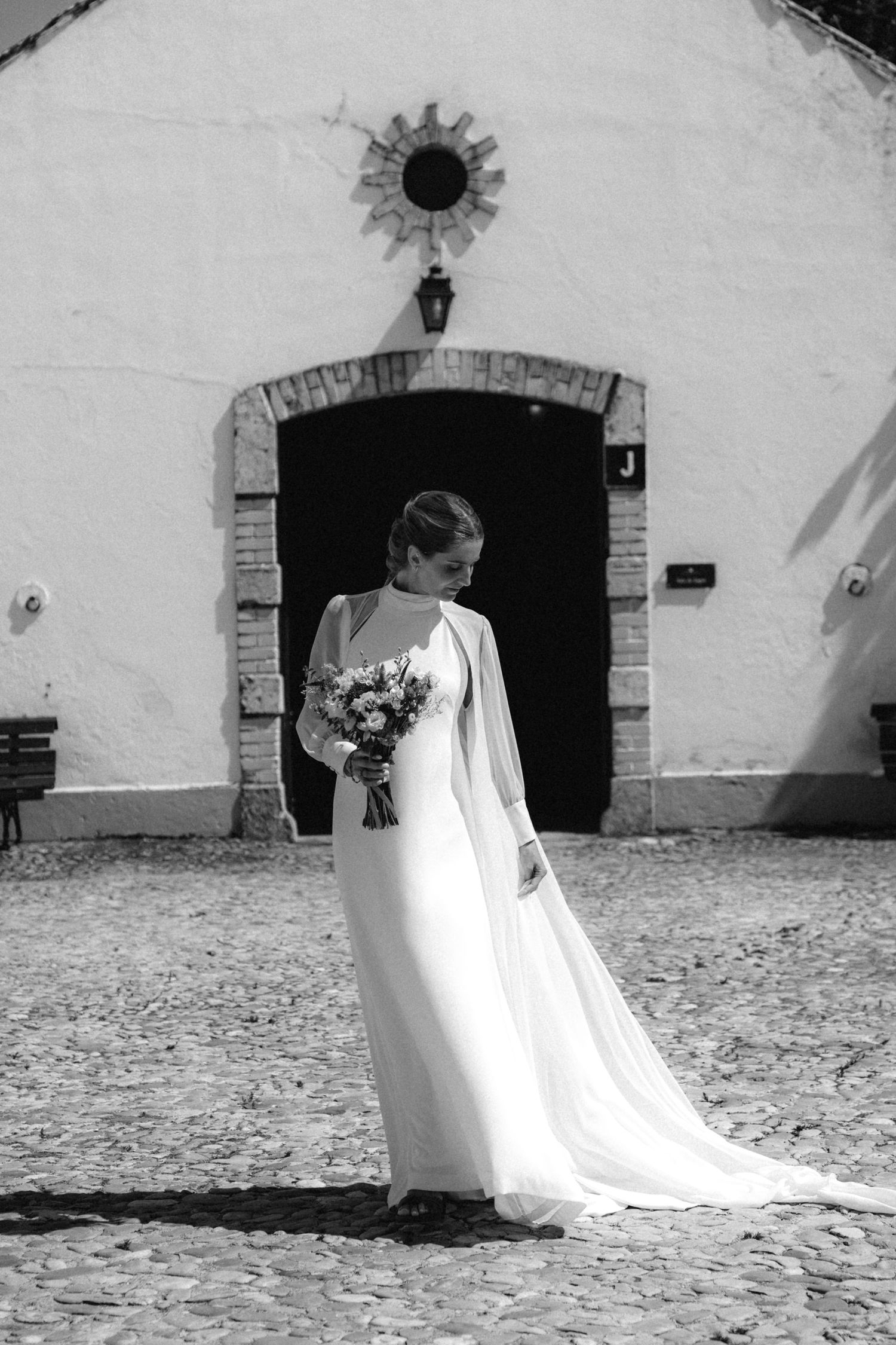 Wedding dress and bouquet silhouetted against white Mediterranean building exterior.
