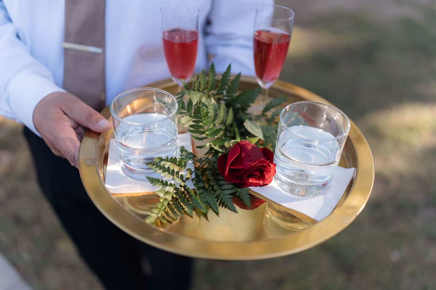 A gold serving tray holds two glasses of rose wine, water glasses, and is decorated with ferns and a red rose.