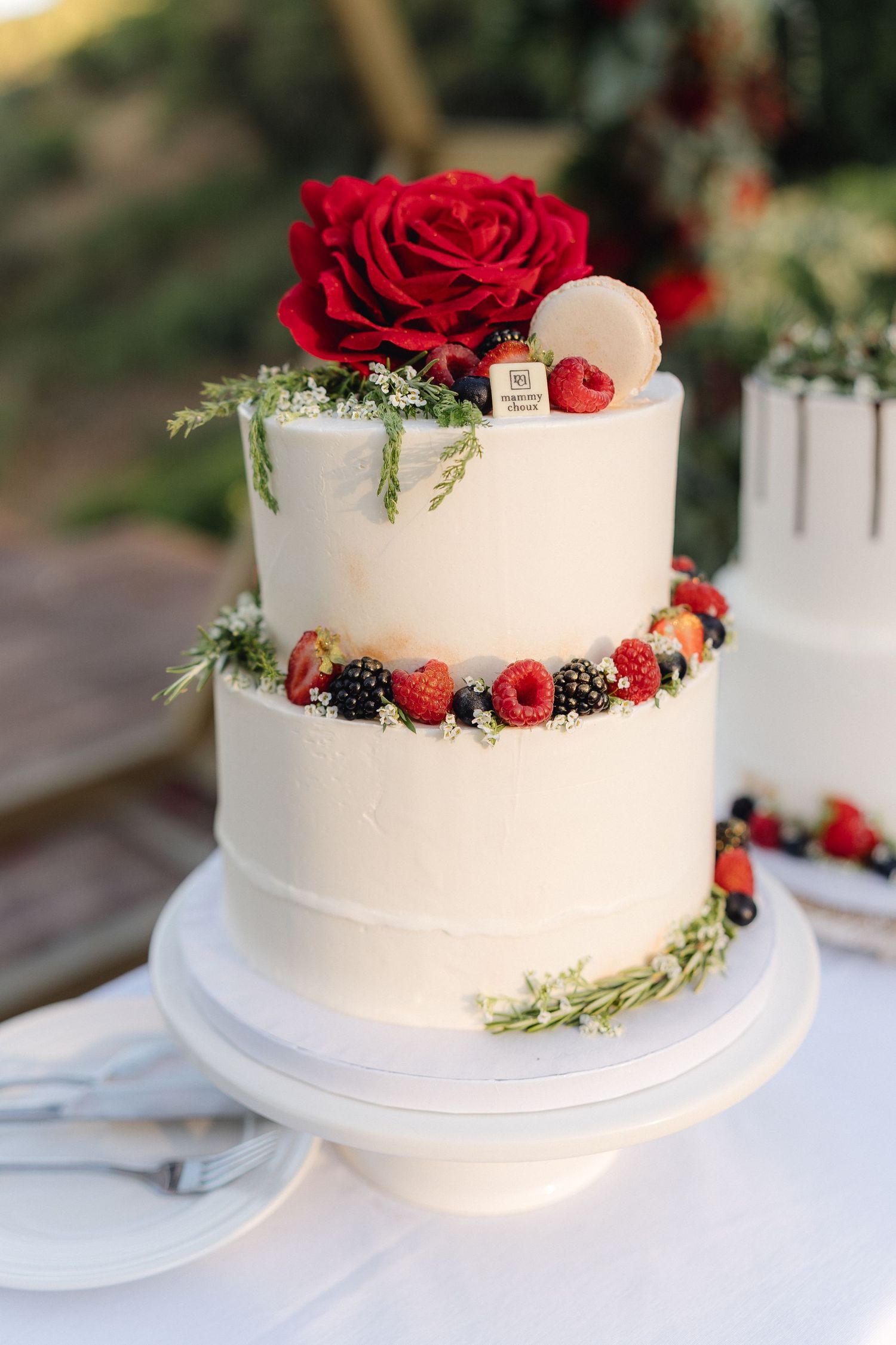 White wedding cake decorated with fresh red roses, berries and greenery on a white cake stand.