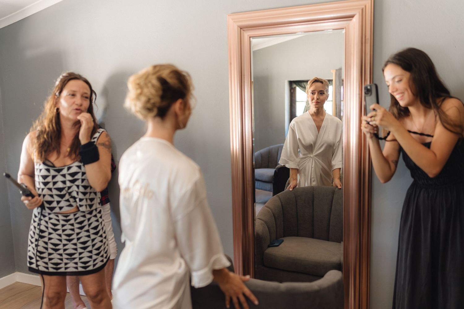 Wedding party getting ready in a room with a large mirror and gray walls.