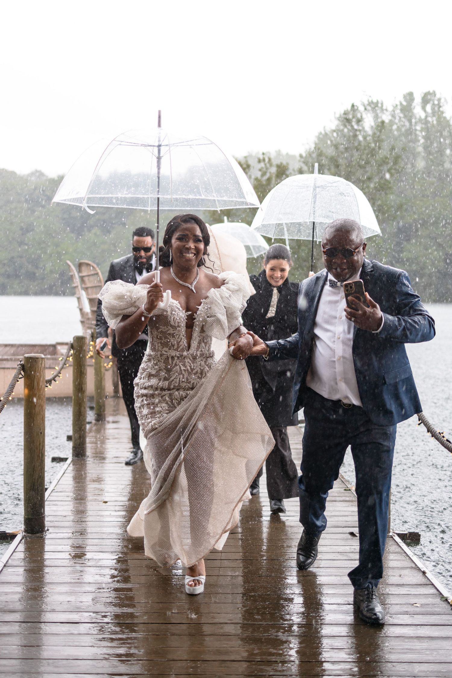 Wedding party running through the rain on a wooden dock while holding umbrellas.
