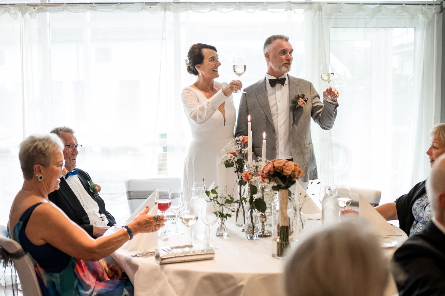 Wedding reception guests raising glasses for toast in bright sunlit room.