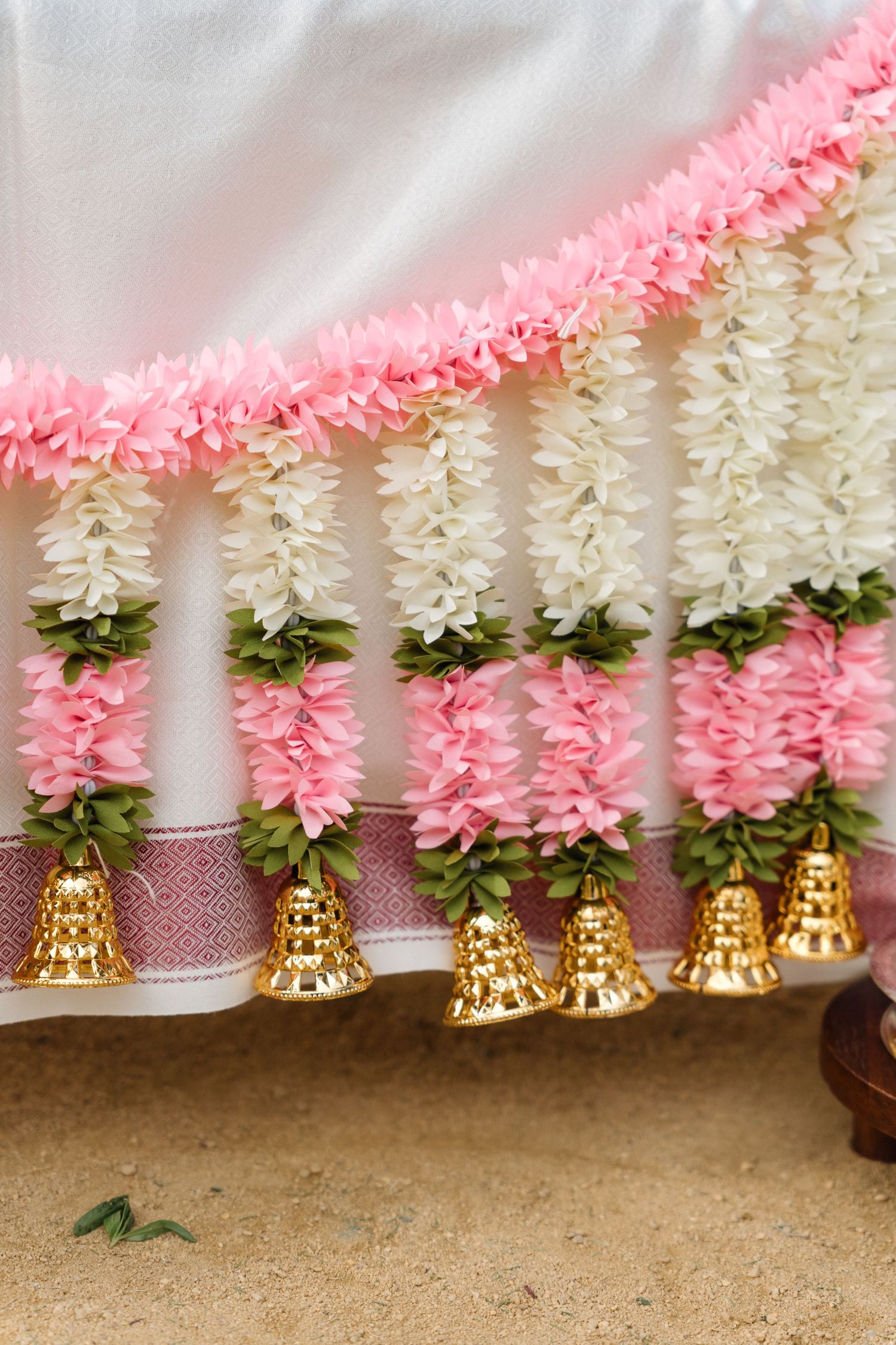 Decorative pink and white flower garlands with golden bells hanging against white backdrop.