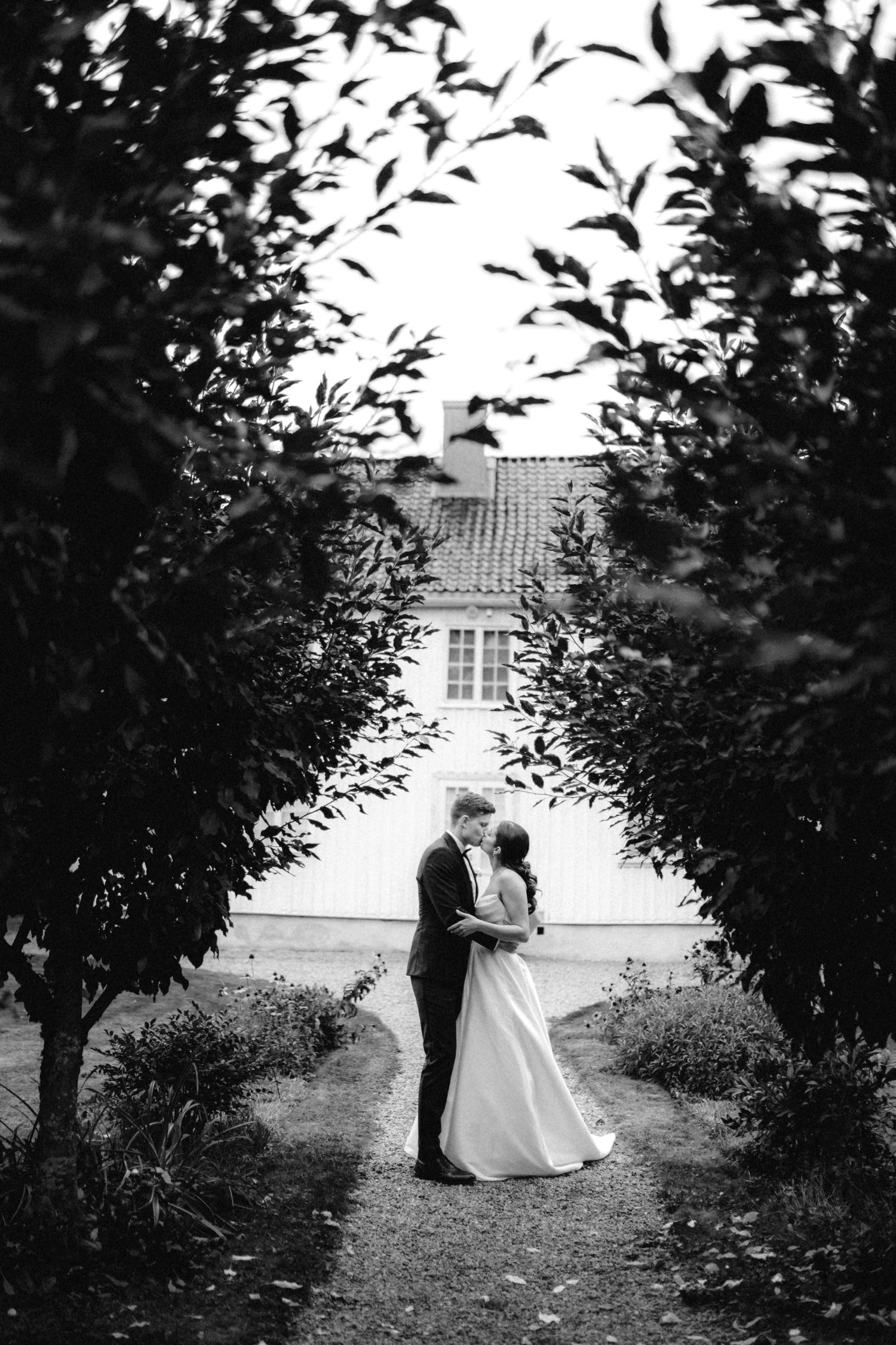 Black and white photo of a bride and groom embracing in a garden path framed by lush trees near a white building.