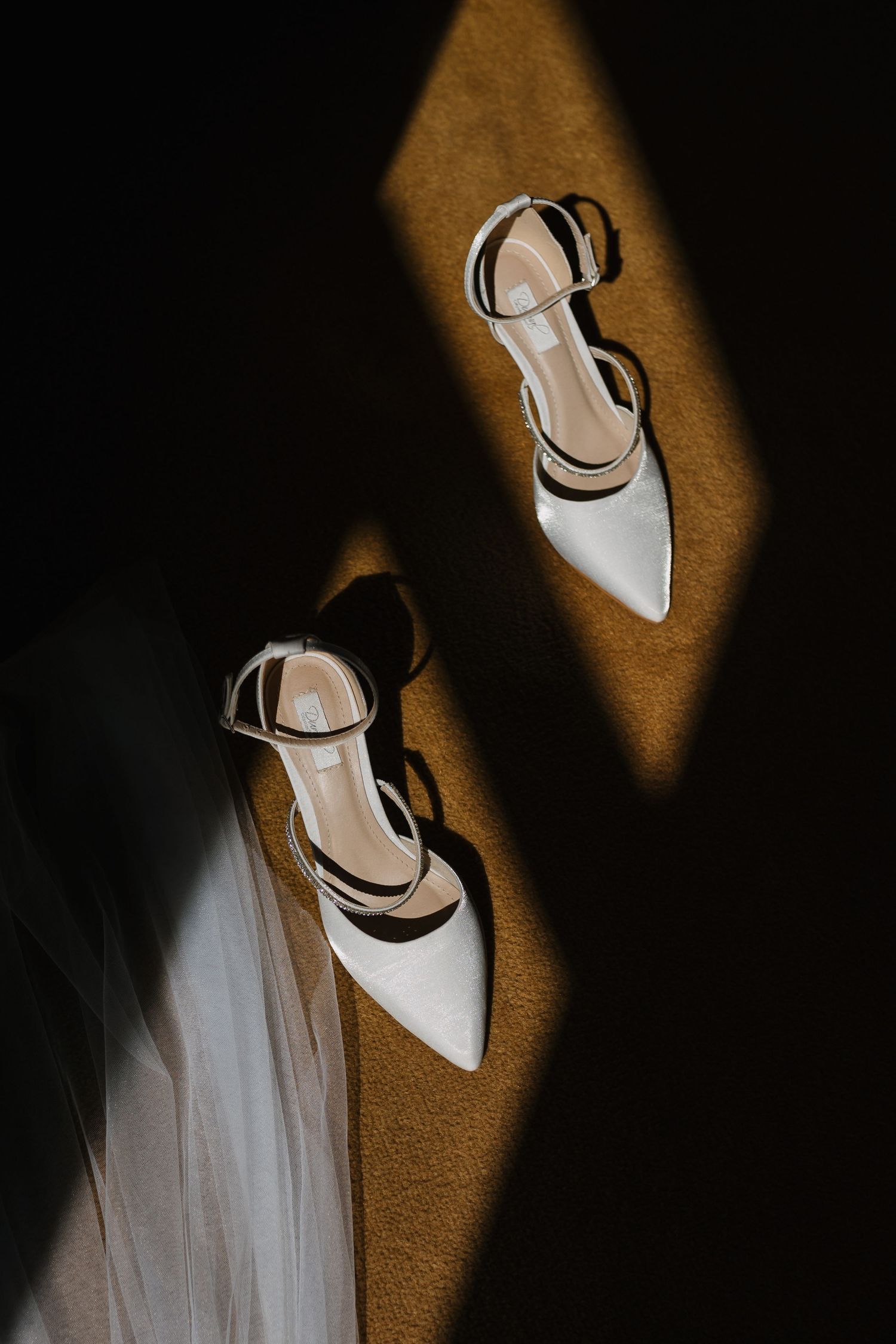 Dramatic lighting on white bridal shoes against dark background creating artistic shadows.