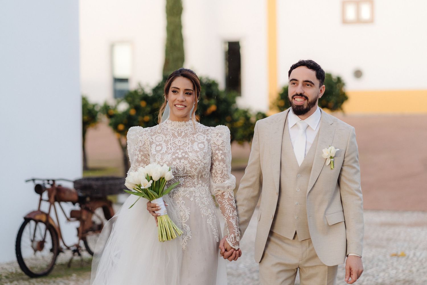 Bride in lace gown holding white bouquet walks hand-in-hand with groom in beige suit at outdoor wedding venue.
