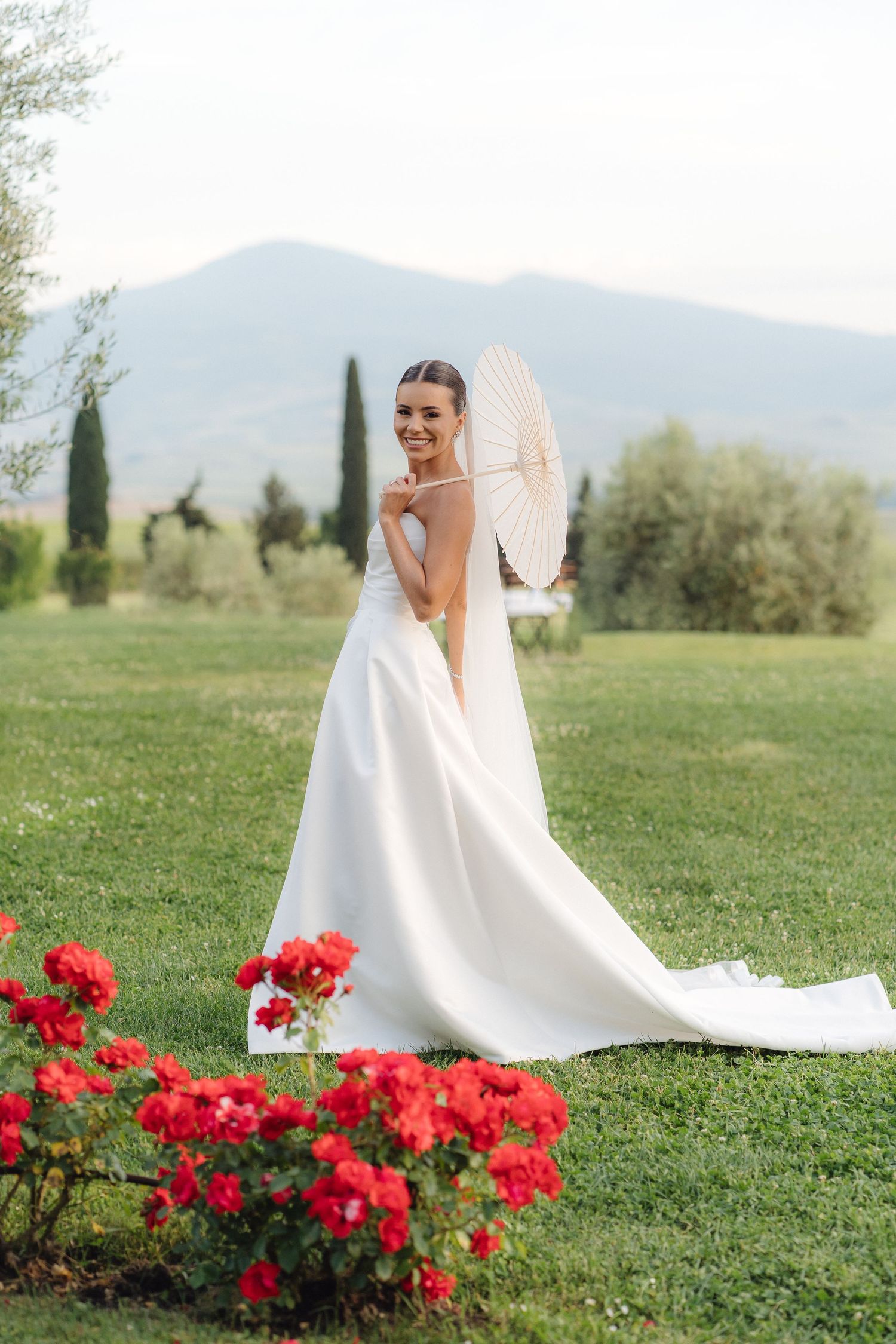 Elegant bride in a white gown holding a parasol standing among red roses with Tuscan hills in the background.