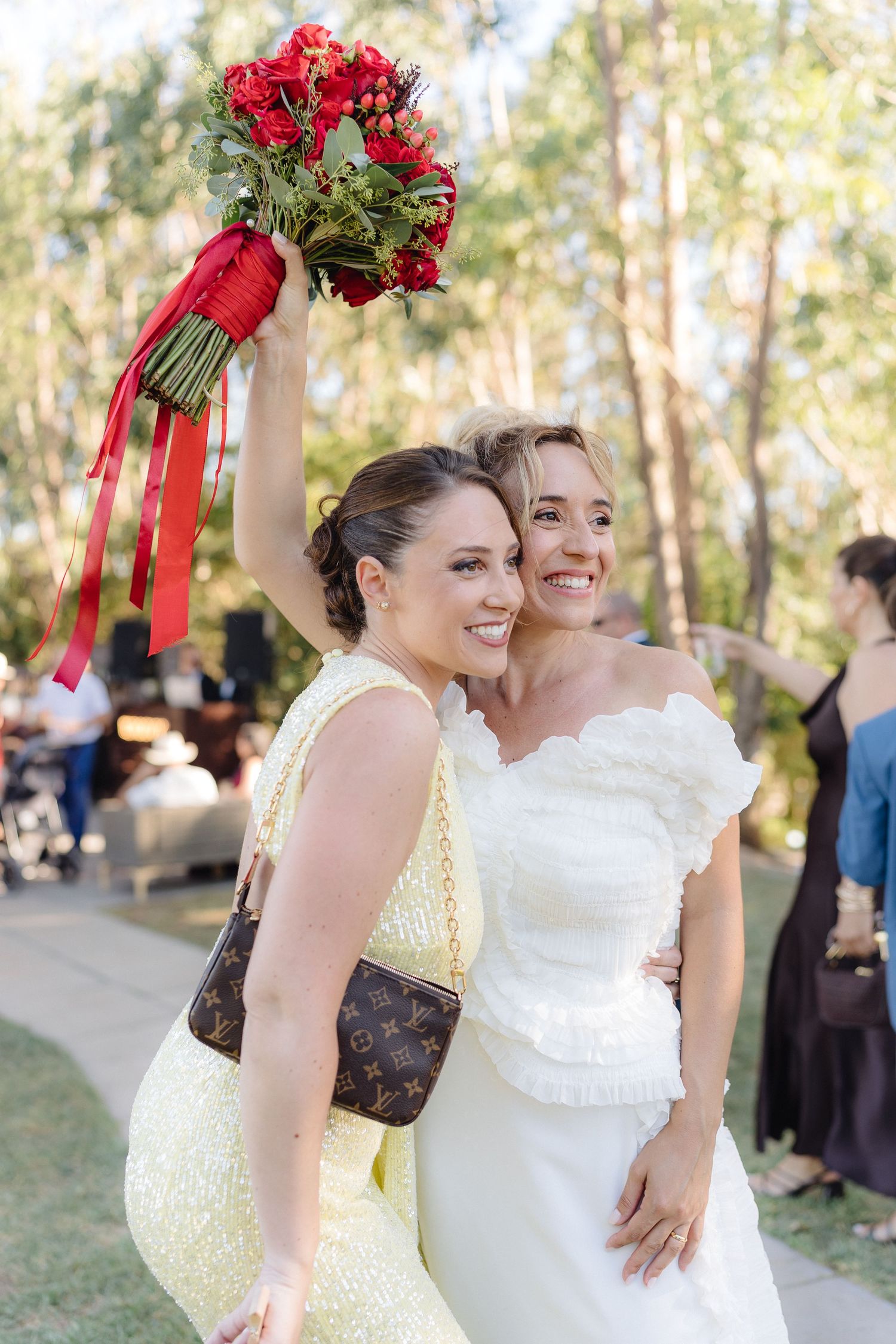 Bride in white dress and bridesmaid in yellow hold vibrant red bouquets outdoors.