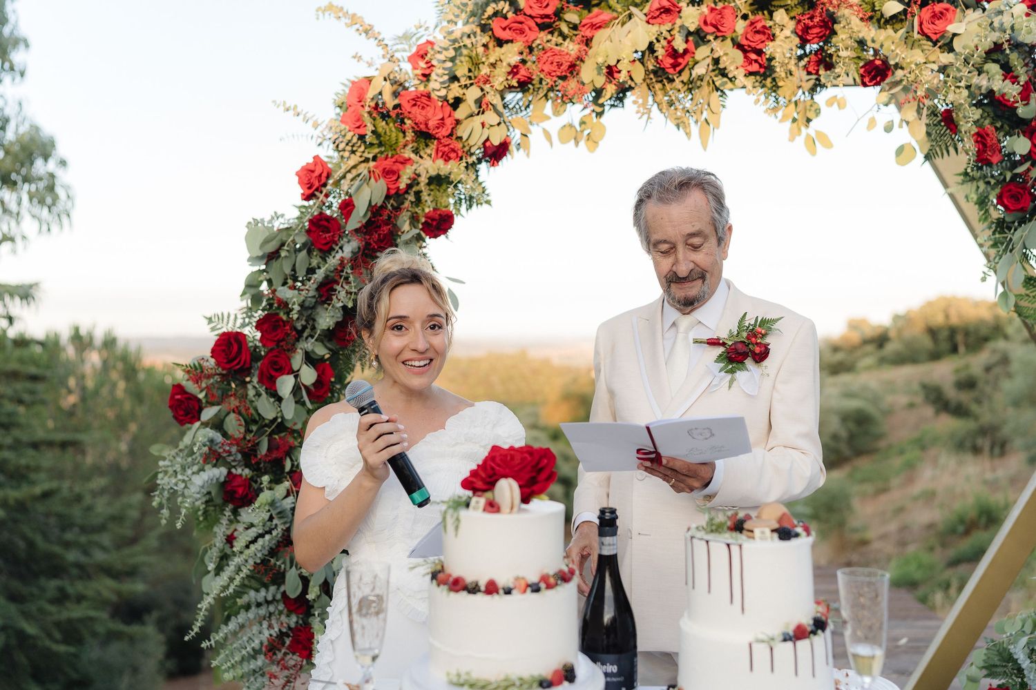 Wedding party toasts with drinks under floral arch decorated with red roses.