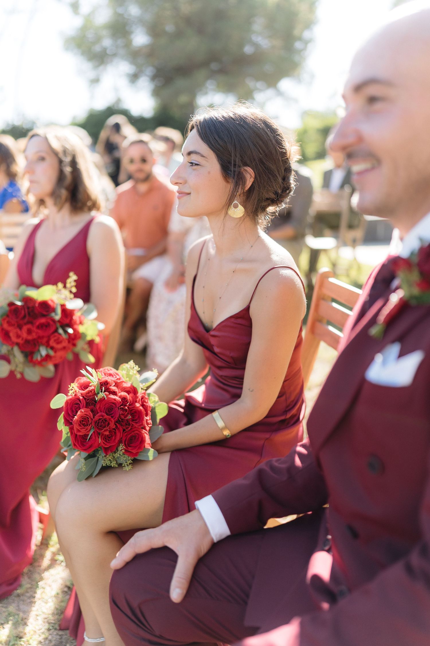 Wedding party members in burgundy dresses hold vibrant red rose bouquets during an outdoor ceremony.