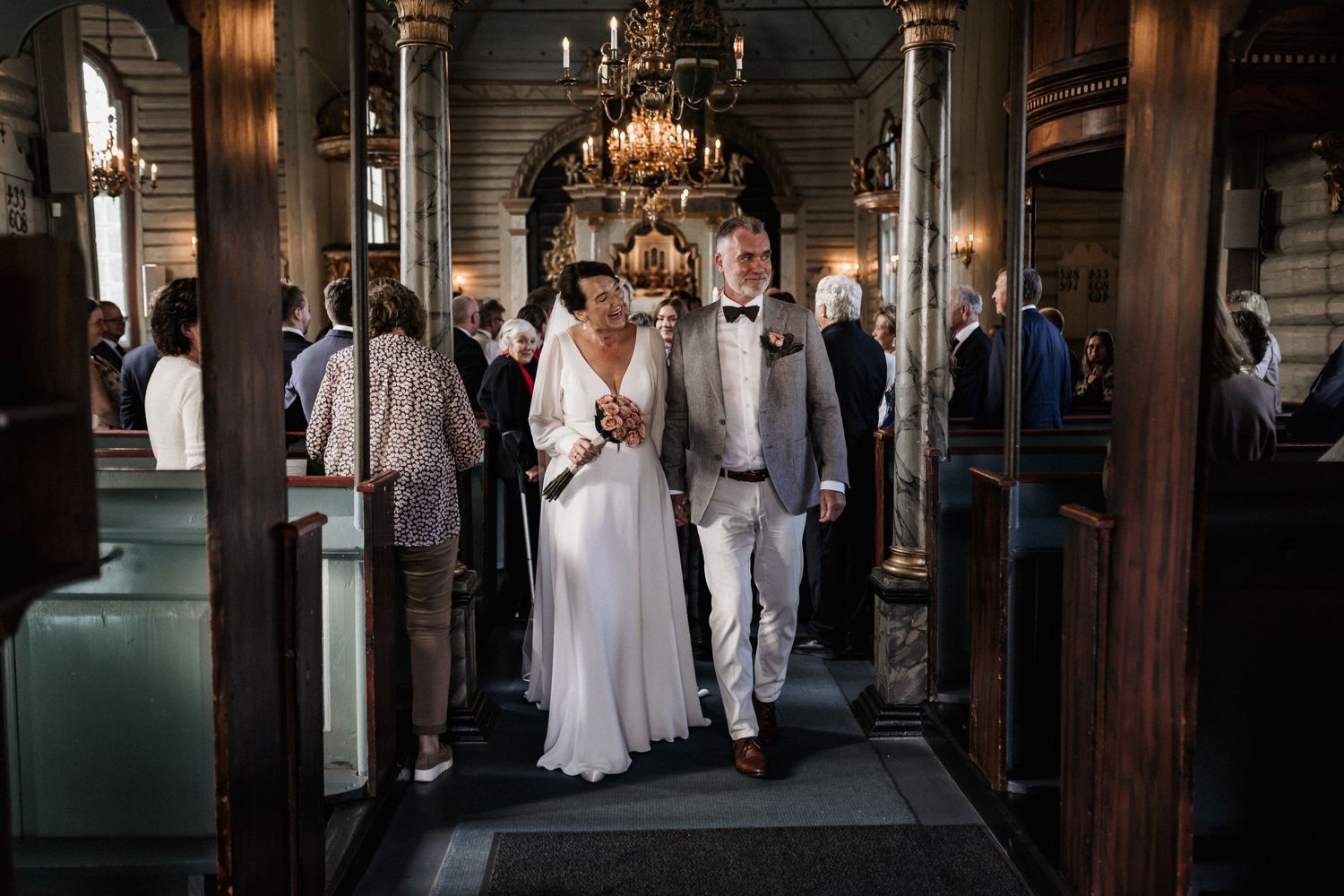 Newlyweds walk down the aisle of a grand church with wooden pillars and chandeliers.
