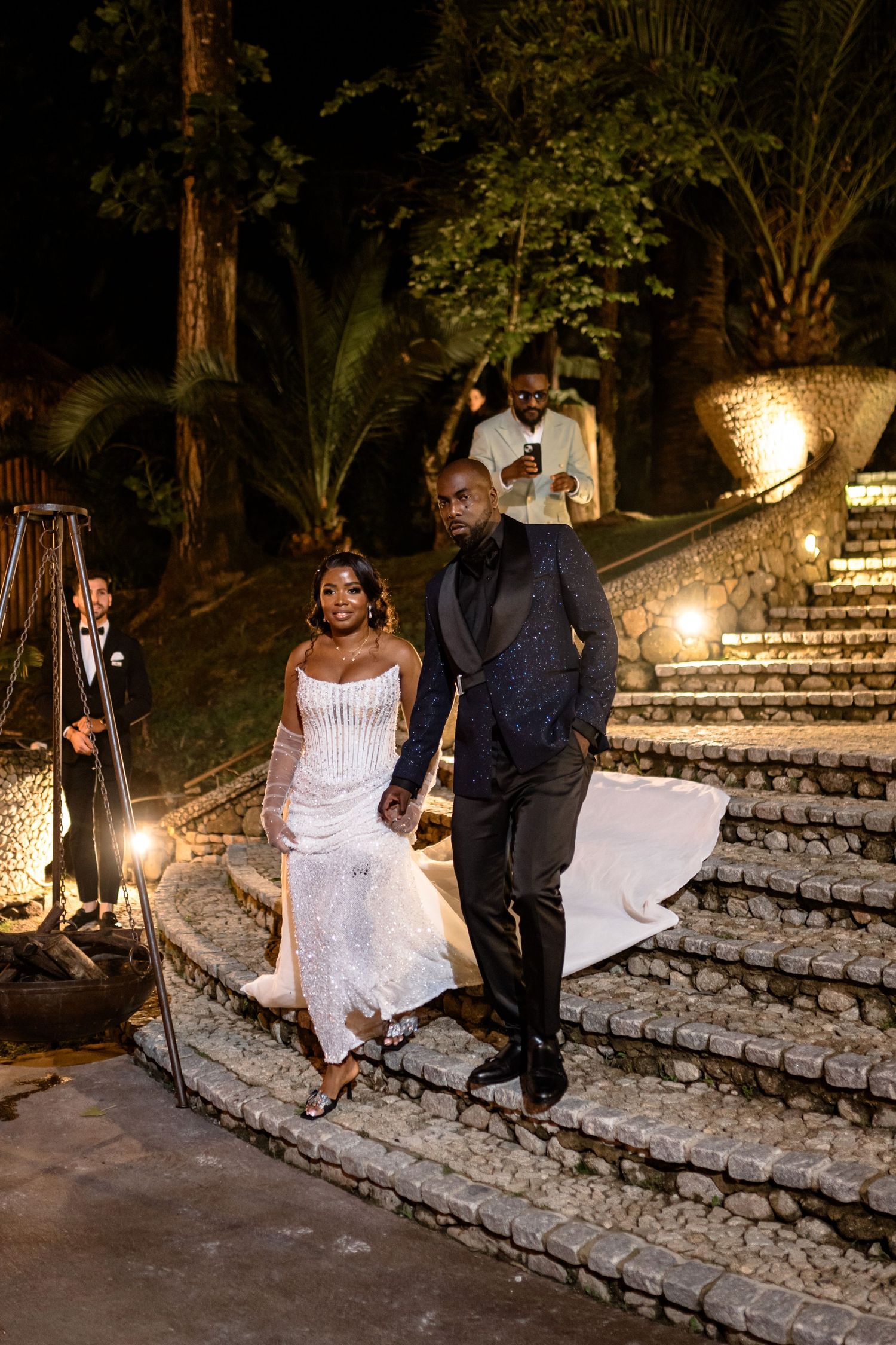 A couple descends illuminated stone steps at an elegant outdoor evening wedding venue with palm trees and ambient lighting.