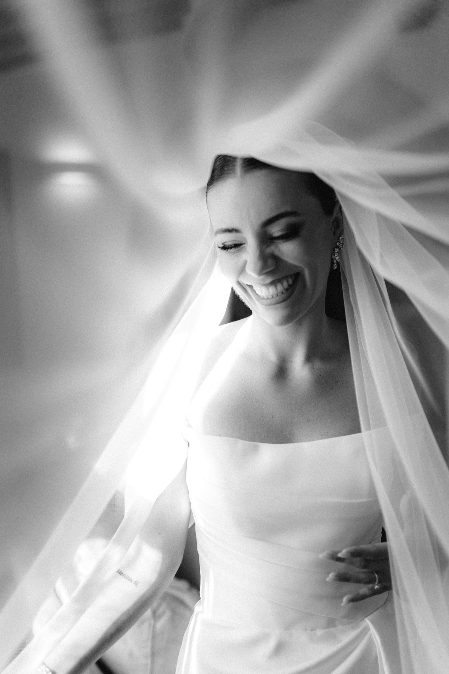 Black and white photo of a radiant bride smiling under her flowing veil with soft light rays surrounding her.