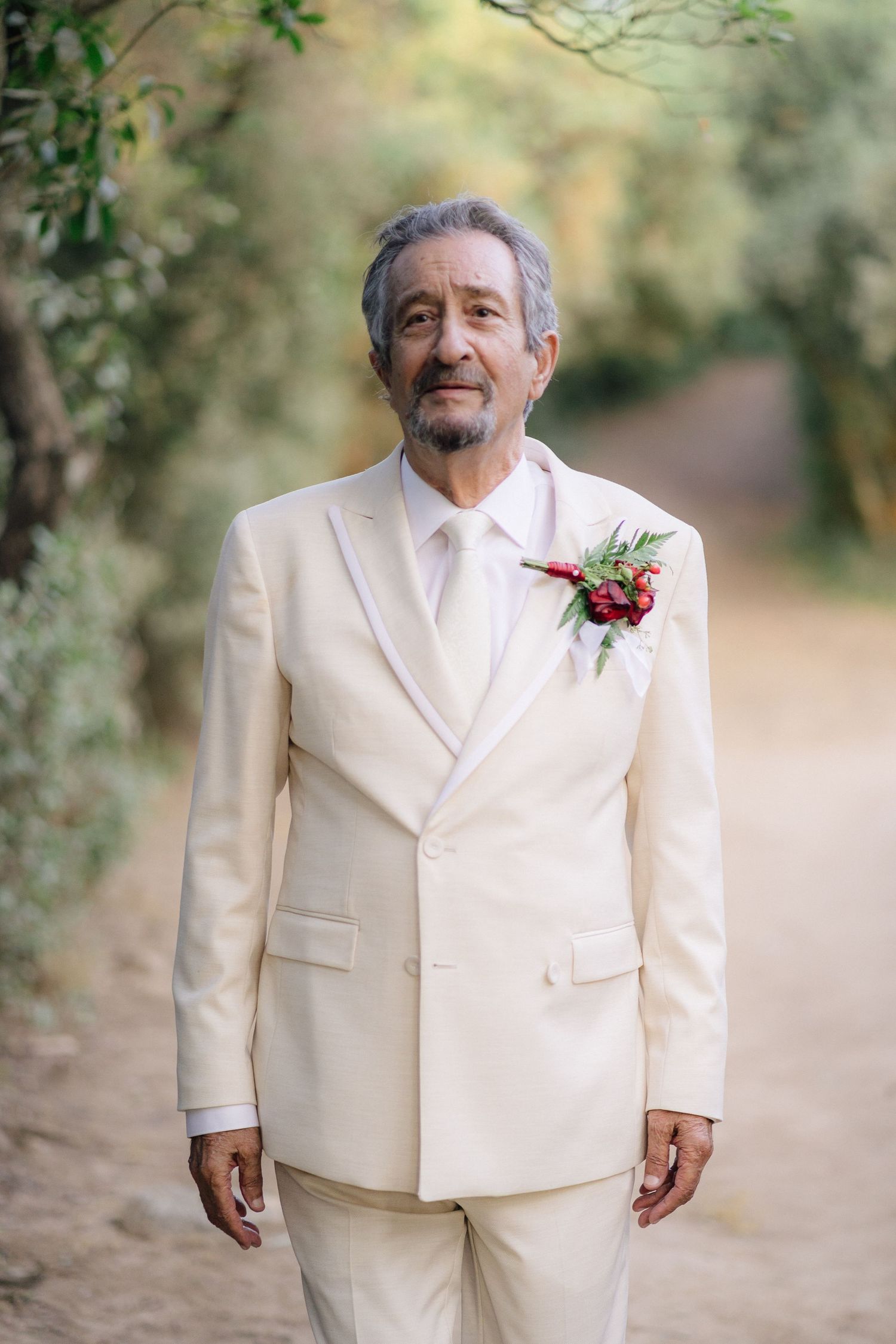 Groom in cream colored suit with red boutonniere poses outdoors.