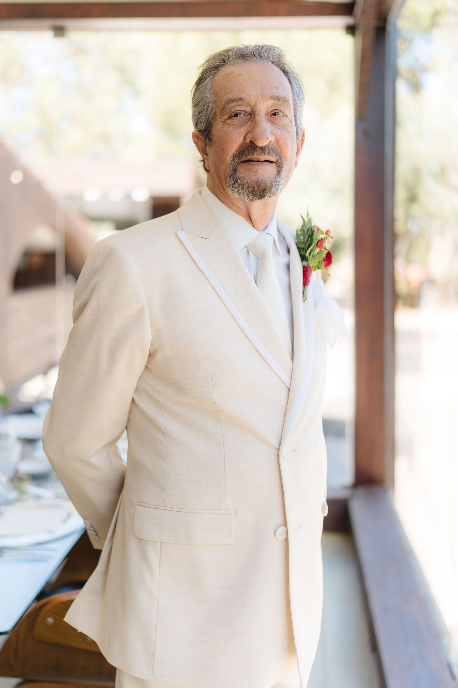 Person in white suit jacket with boutonniere smiling warmly in natural light.