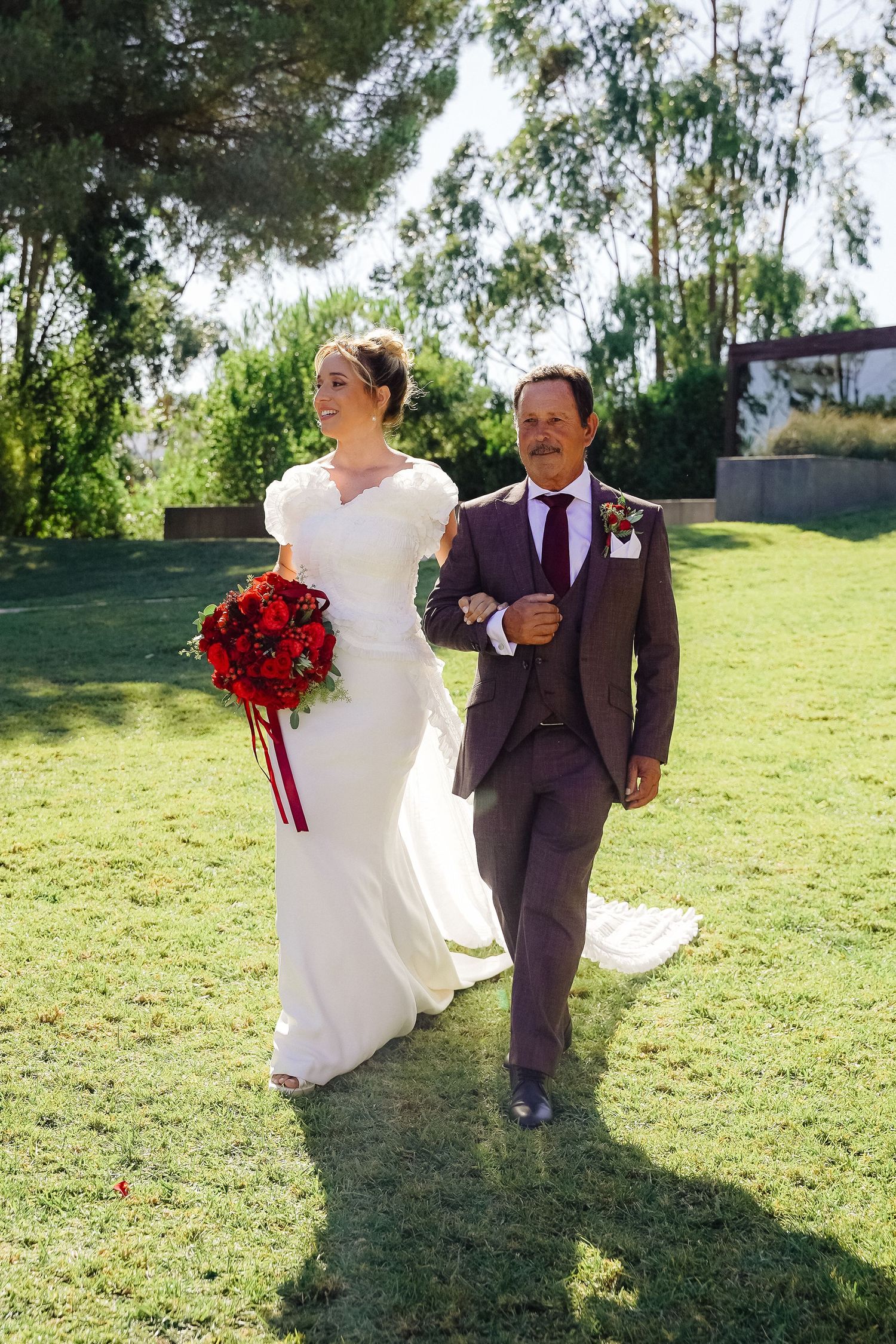 A couple in wedding attire strolls across grass with a stunning red bouquet.