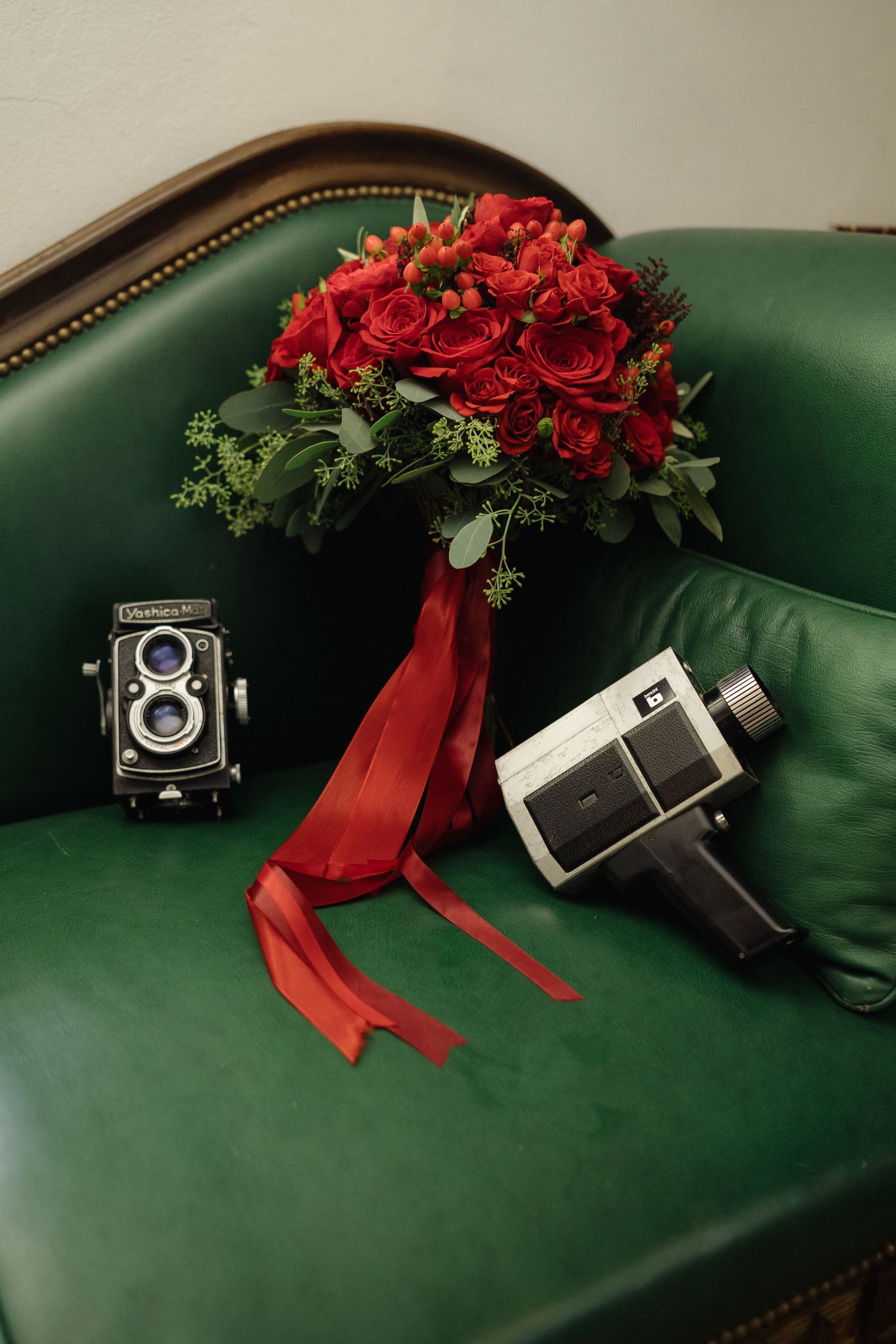 Red rose bouquet with ribbons and vintage cameras arranged on green leather furniture.