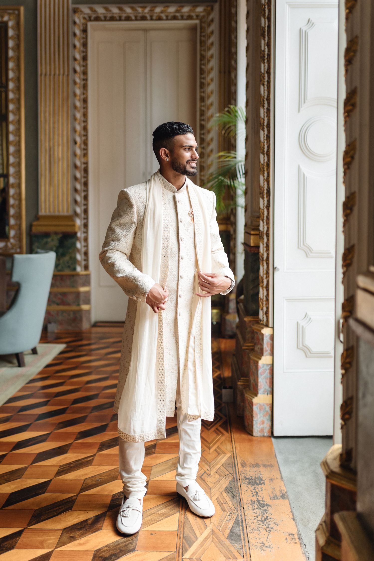 Person in traditional cream Indian wedding attire standing in ornate room with patterned floor.