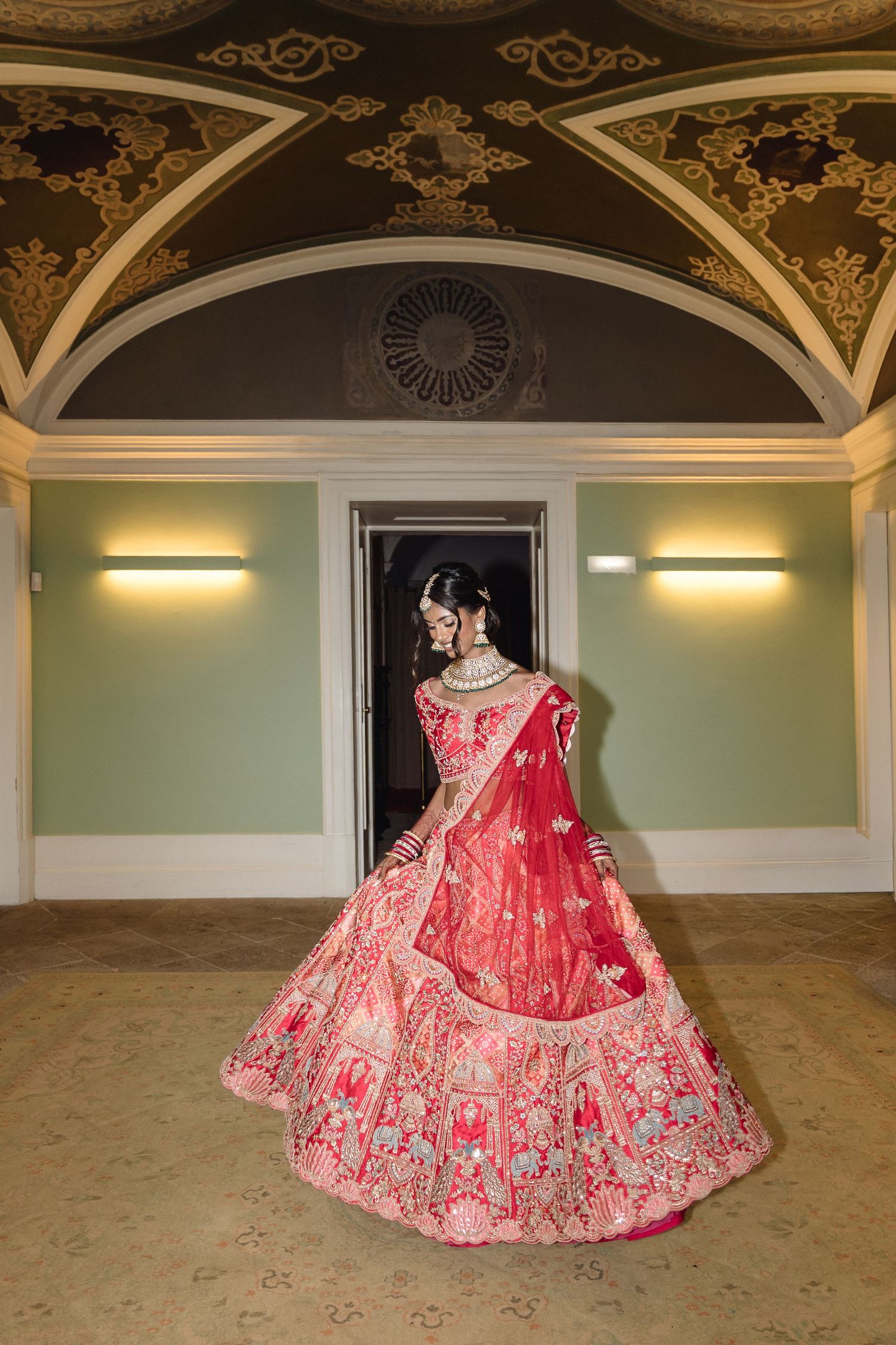Elaborate red bridal lehenga photographed under ornate gilded ceiling in grand ballroom.