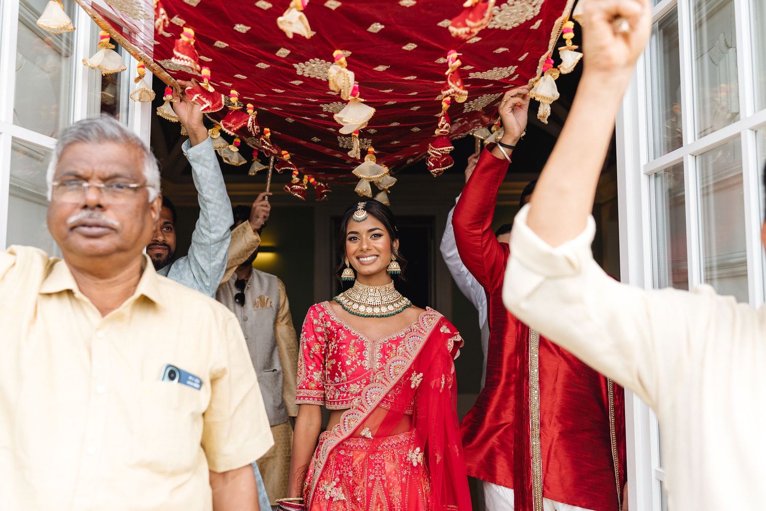 Wedding party members in traditional Indian attire celebrate under red and gold decorative ceiling.