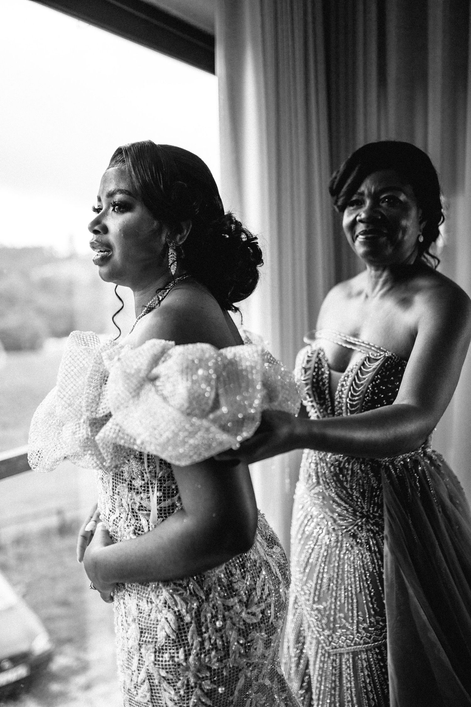 Two women in elegant beaded wedding gowns share an intimate moment during bridal preparations.