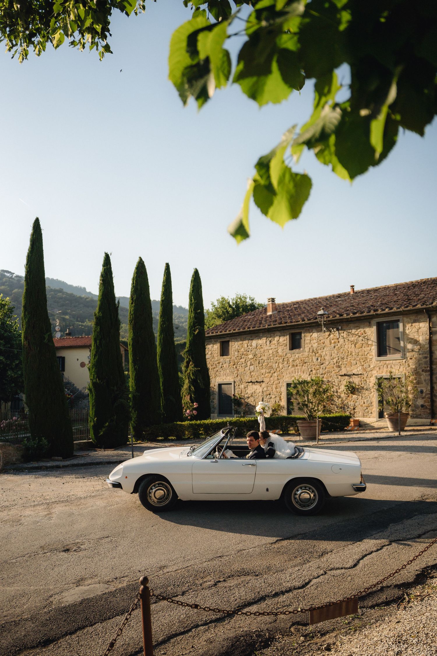 Couple riding in a vintage white convertible car outside a rustic stone villa with tall cypress trees in Tuscany.