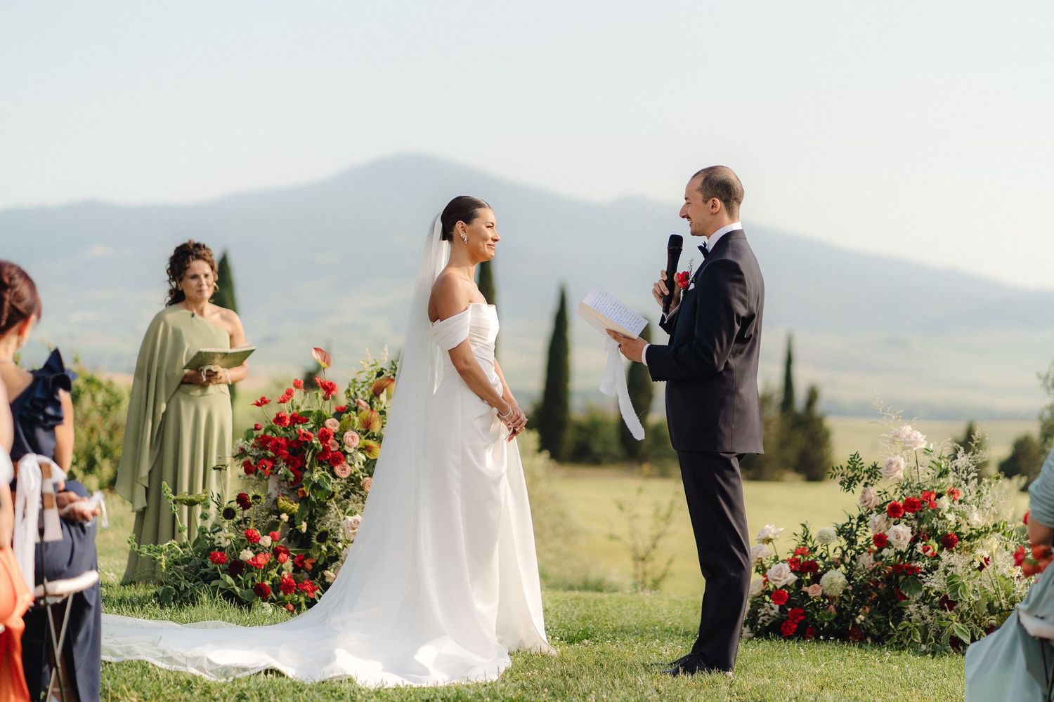 Bride and groom exchange vows outdoors with colorful floral arrangements and Tuscan hills in the background.