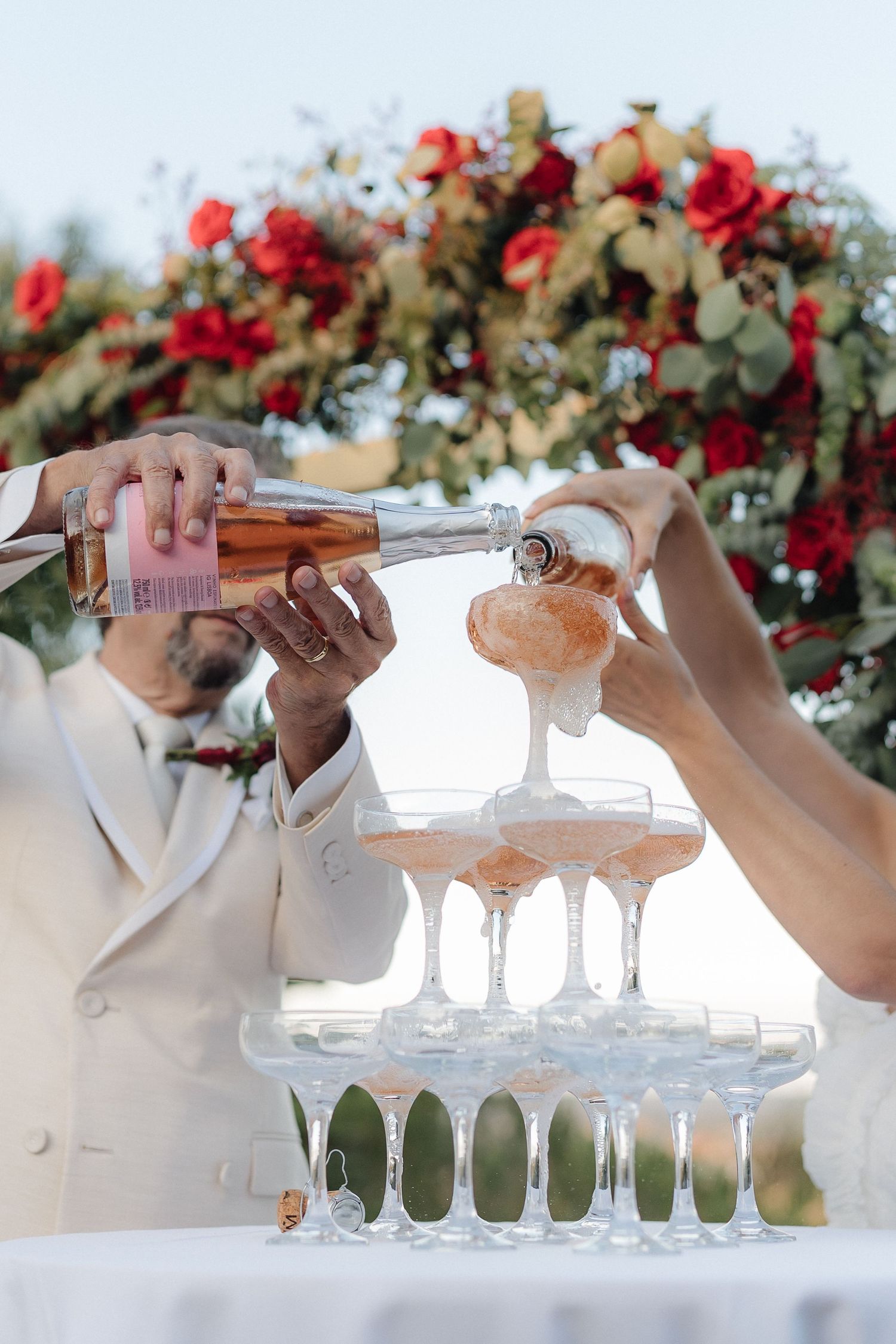 Hands pouring champagne into tower of glasses at outdoor wedding reception.