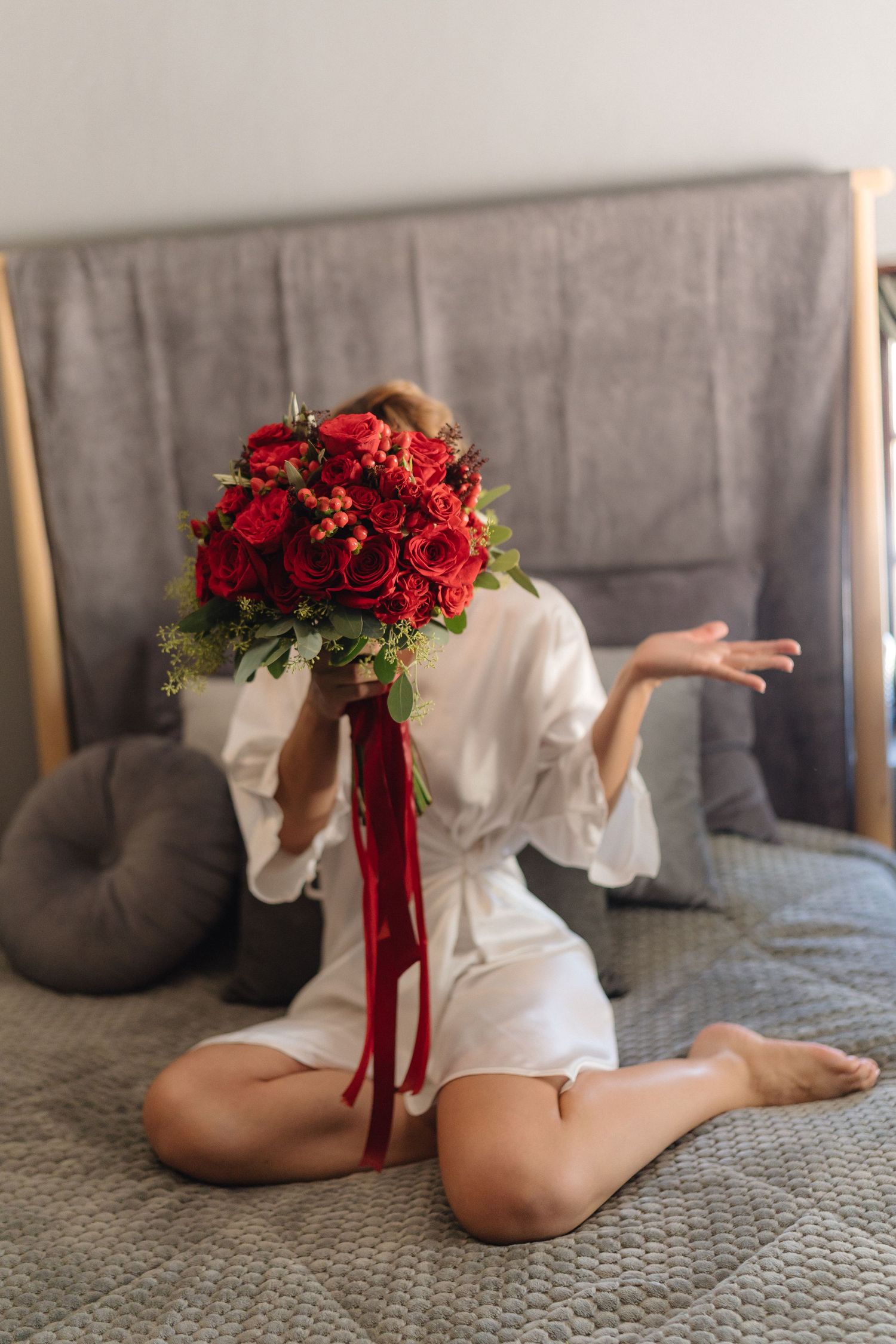 White-robed figure on grey bedding holding vibrant red rose arrangement.