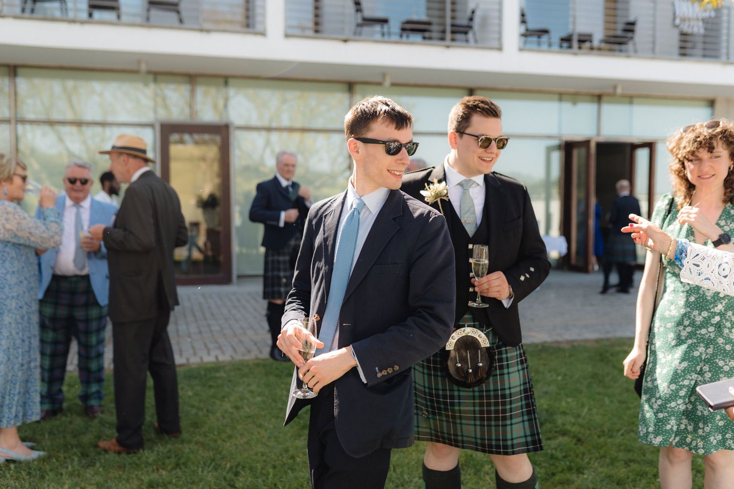 Guests in formal attire including kilts mingle on a lawn during an outdoor event.