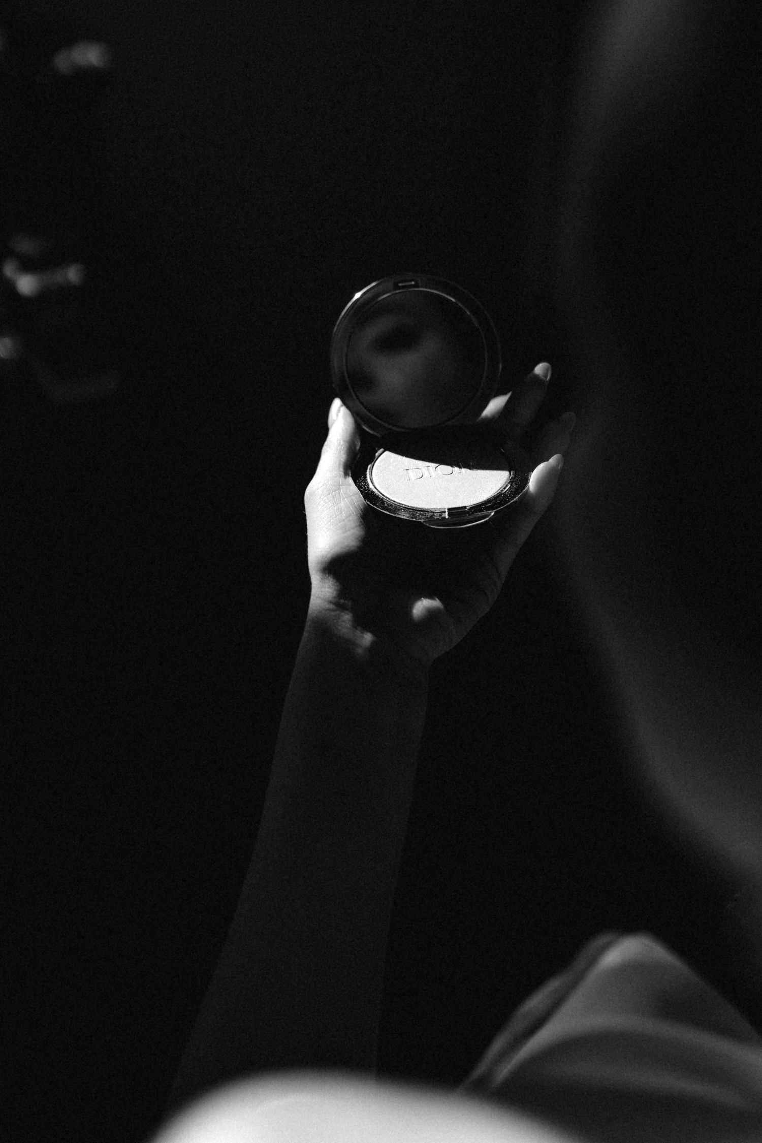 Black and white photo of a hand holding an open compact mirror in dim lighting.