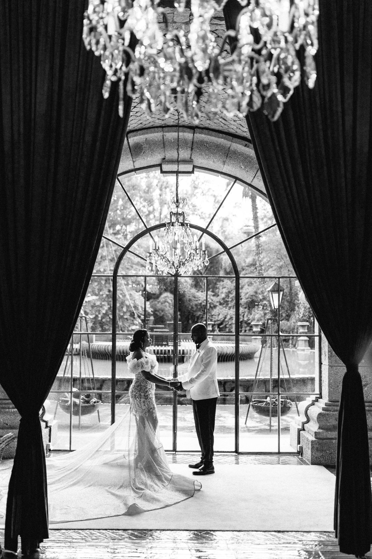 Black and white photo of couple under crystal chandelier by arched window.