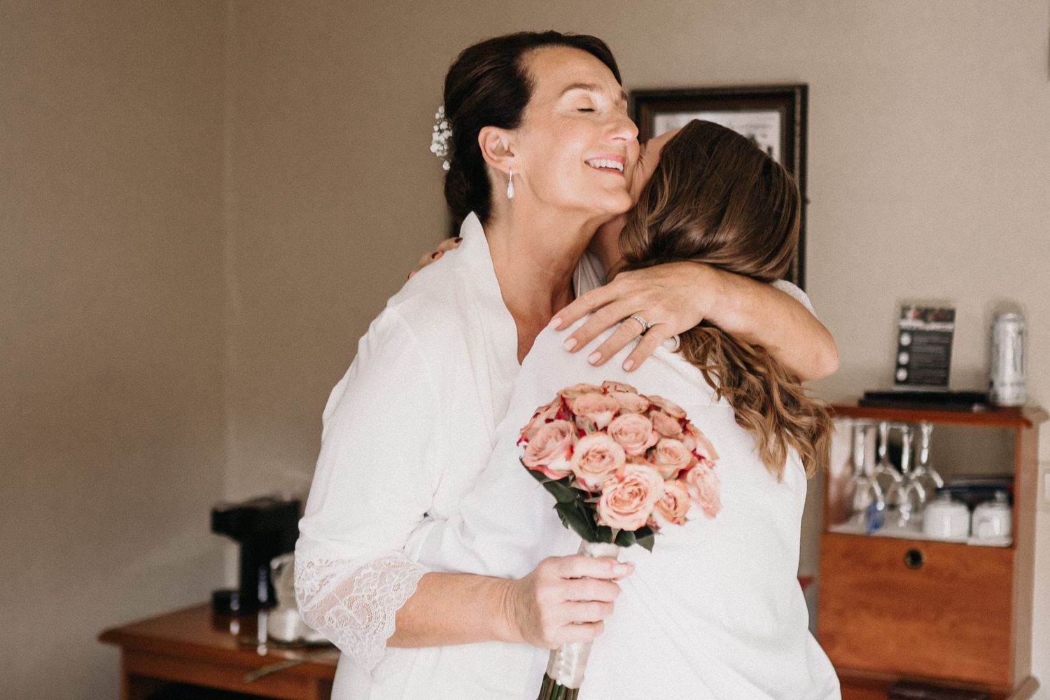 A couple shares a tender embrace during a joyful wedding celebration in a light-filled room.