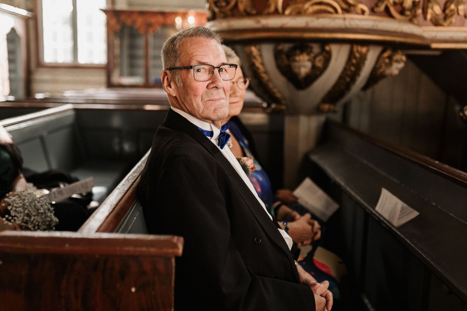 A well-dressed guest sits in an ornate wooden church pew during a wedding ceremony.