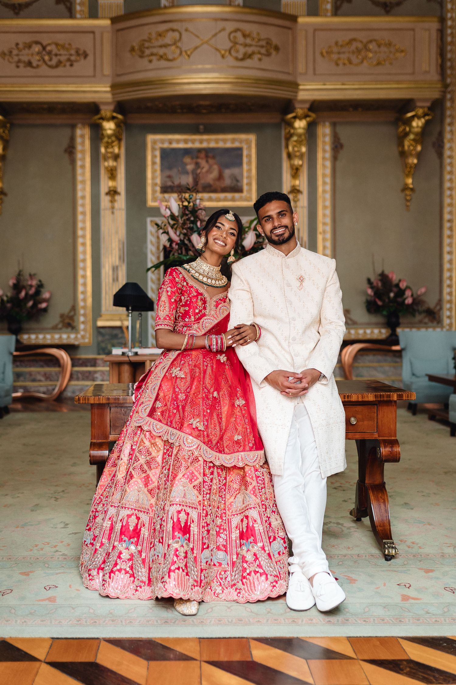 Elegant couple in traditional Indian wedding attire seated in an ornate ballroom with gilded decorations.