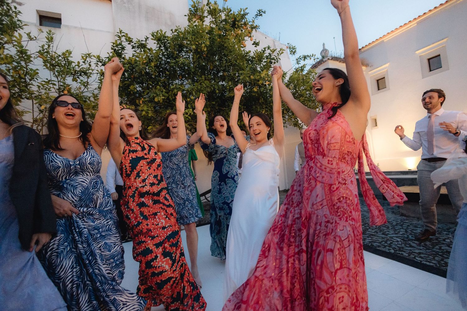 Party guests dancing with raised arms in floral dresses at a sunset celebration.