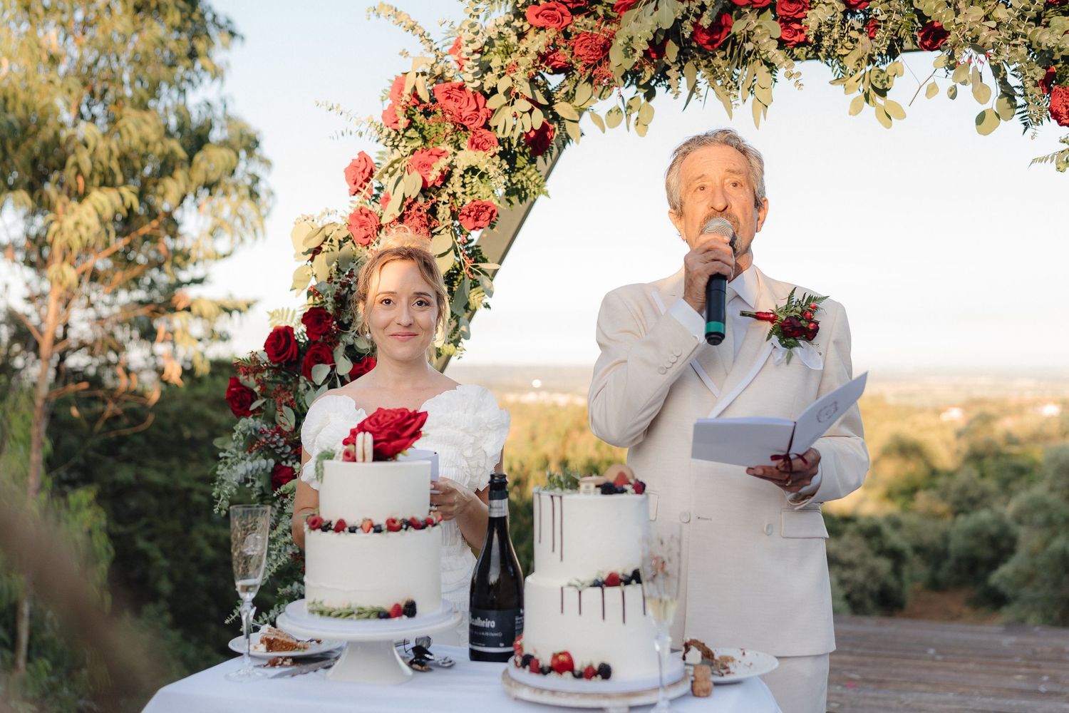 Elegant wedding cake display with red floral decorations and champagne on white tablecloth.